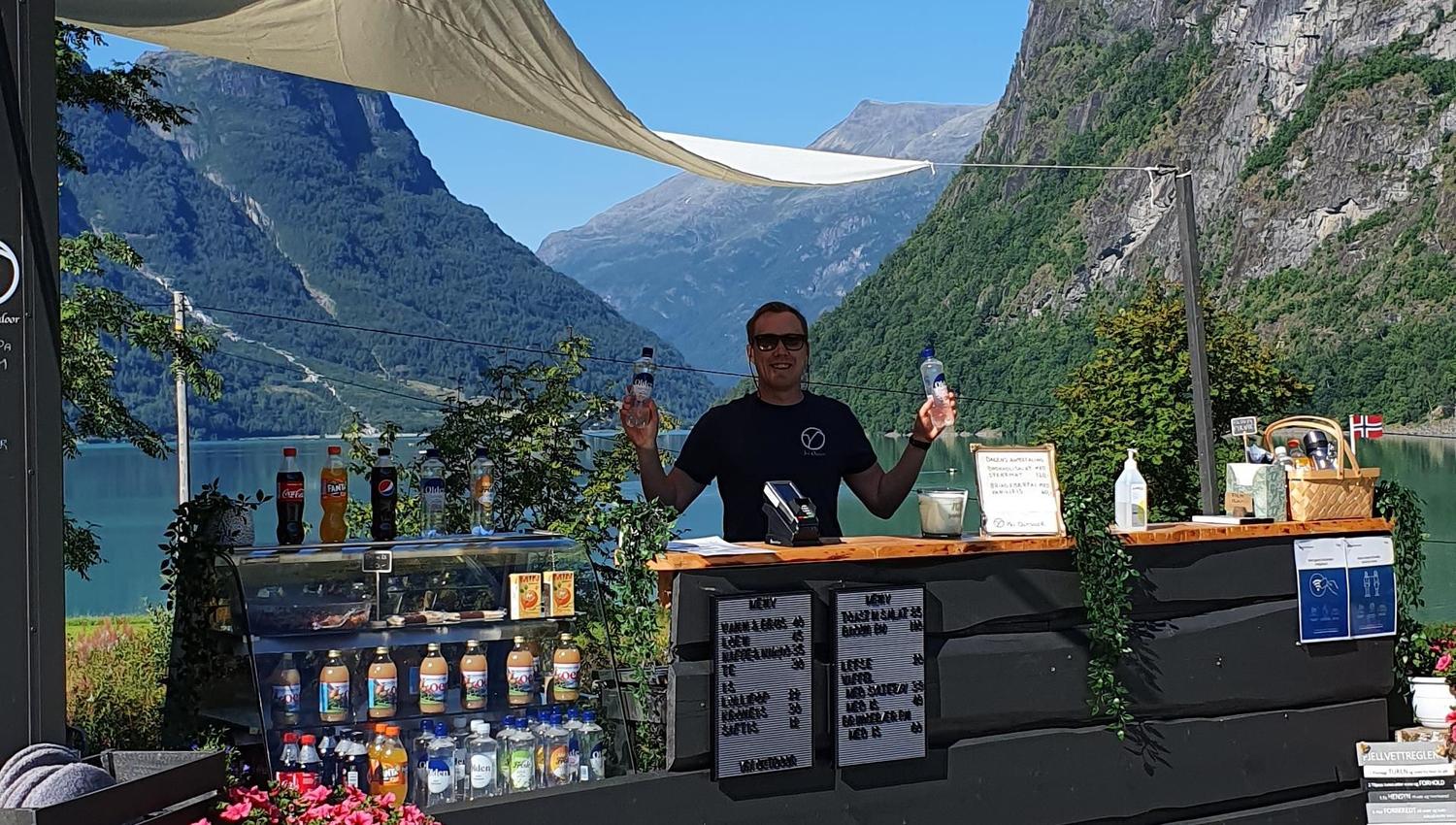 Outdoor drink kiosk with a person behind the counter, bottles and flowers, set by a lake with steep mountains.