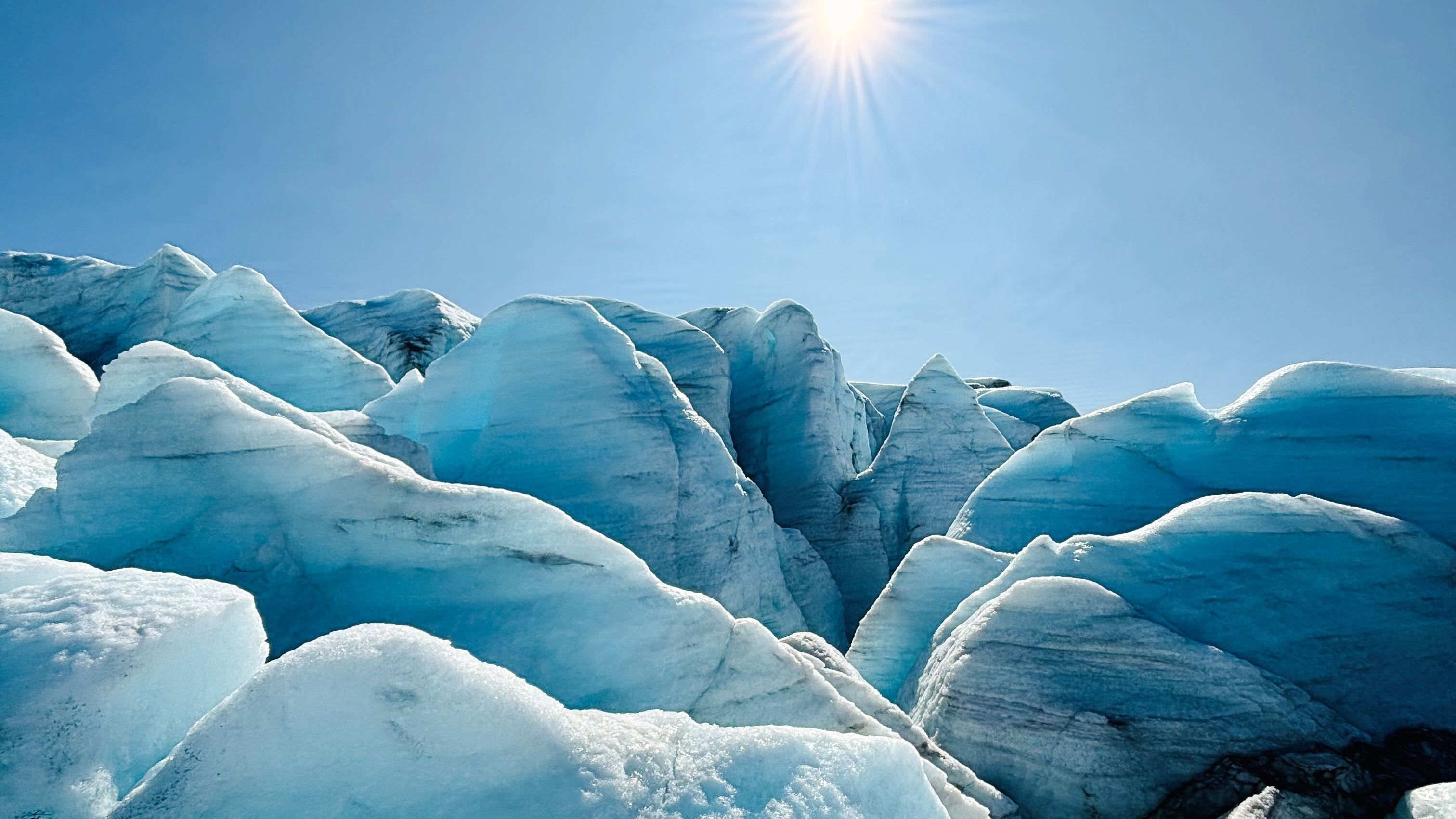 Blue-white glacier ice ridges and crevasses under a clear blue sky, with a bright sun overhead..