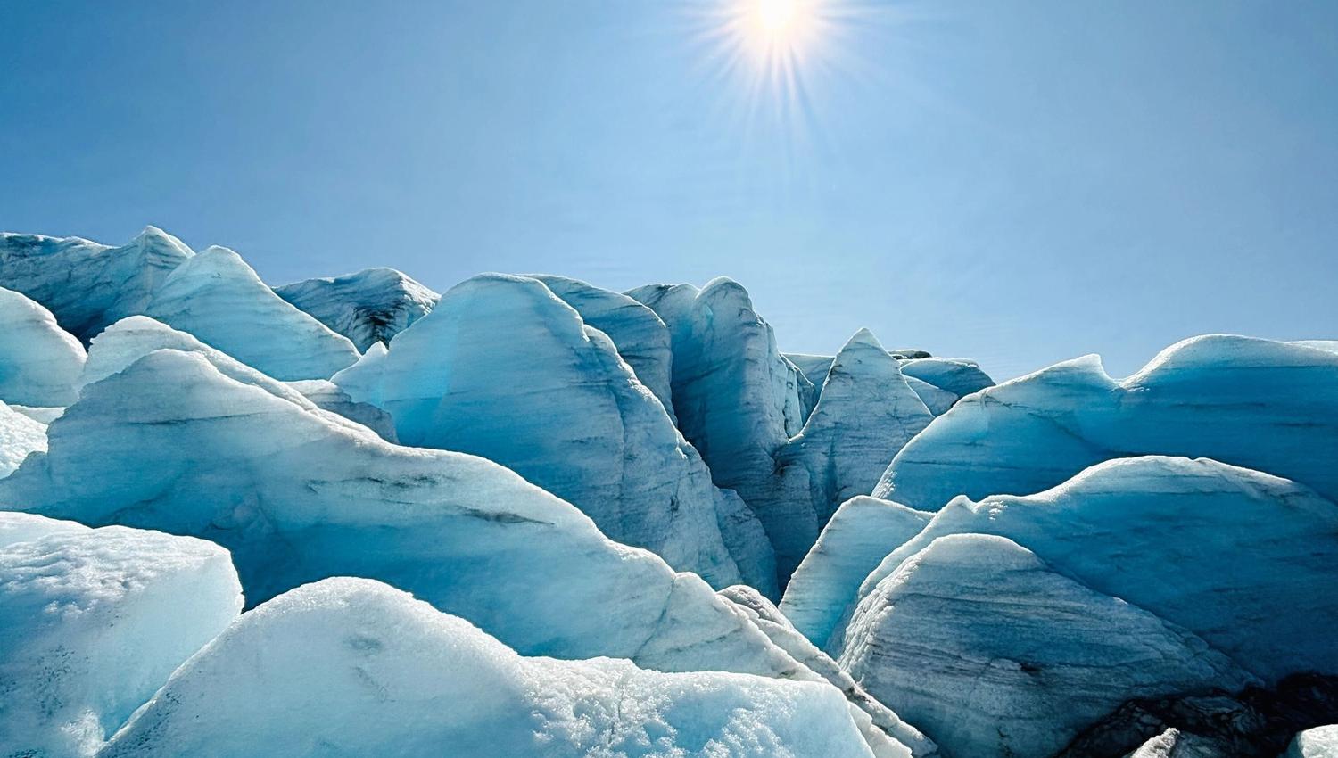 Blue-white glacier ice ridges and crevasses under a clear blue sky, with a bright sun overhead..