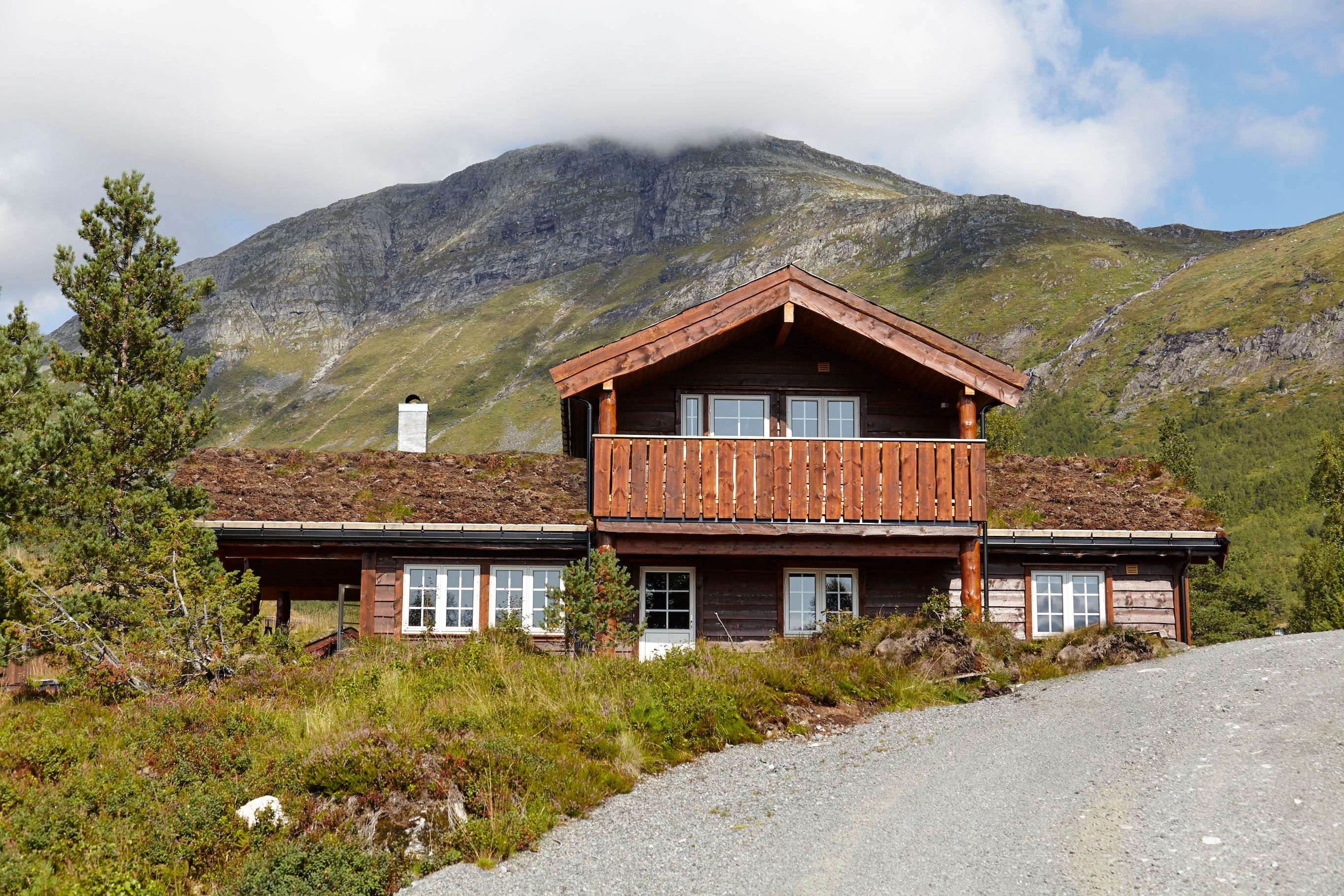 Brun trehytte med balkong og torvtak framfor grøne fjell, med grusveg i forgrunnen og skyer over toppen..