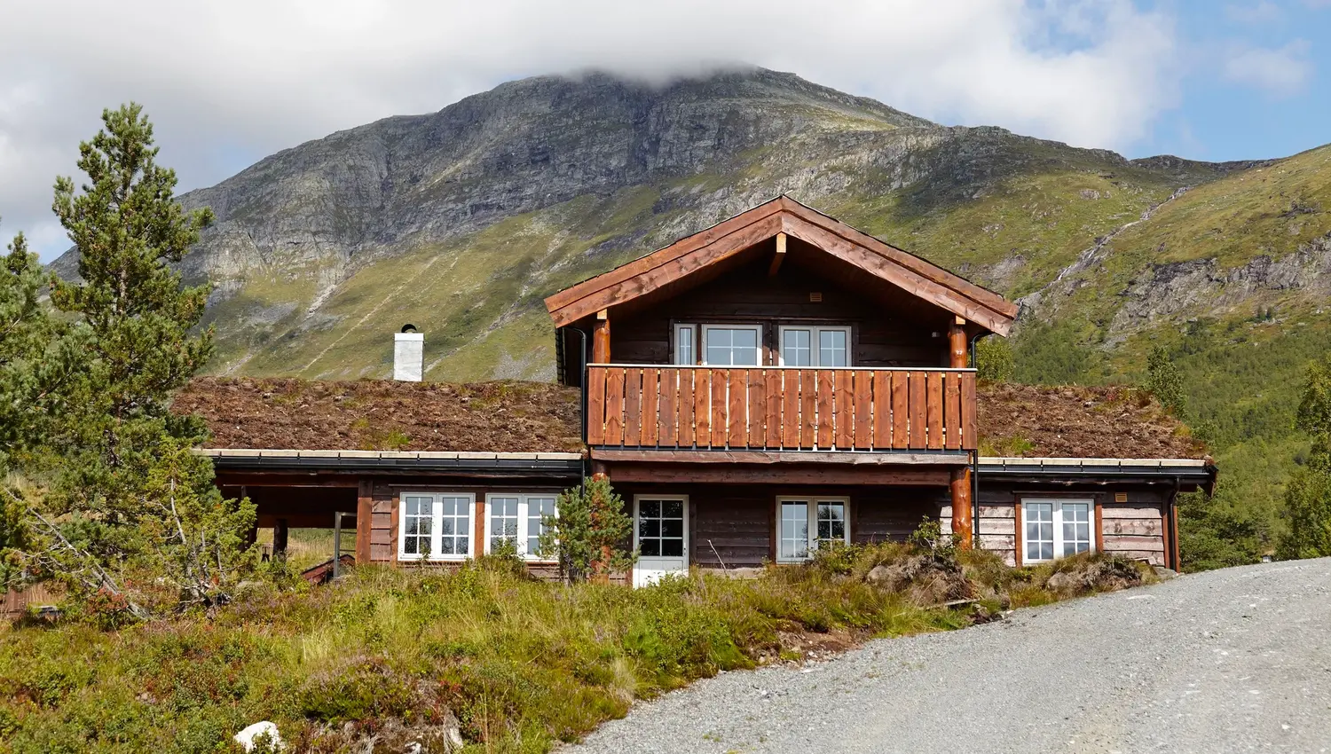 Brun trehytte med balkong og torvtak framfor grøne fjell, med grusveg i forgrunnen og skyer over toppen..