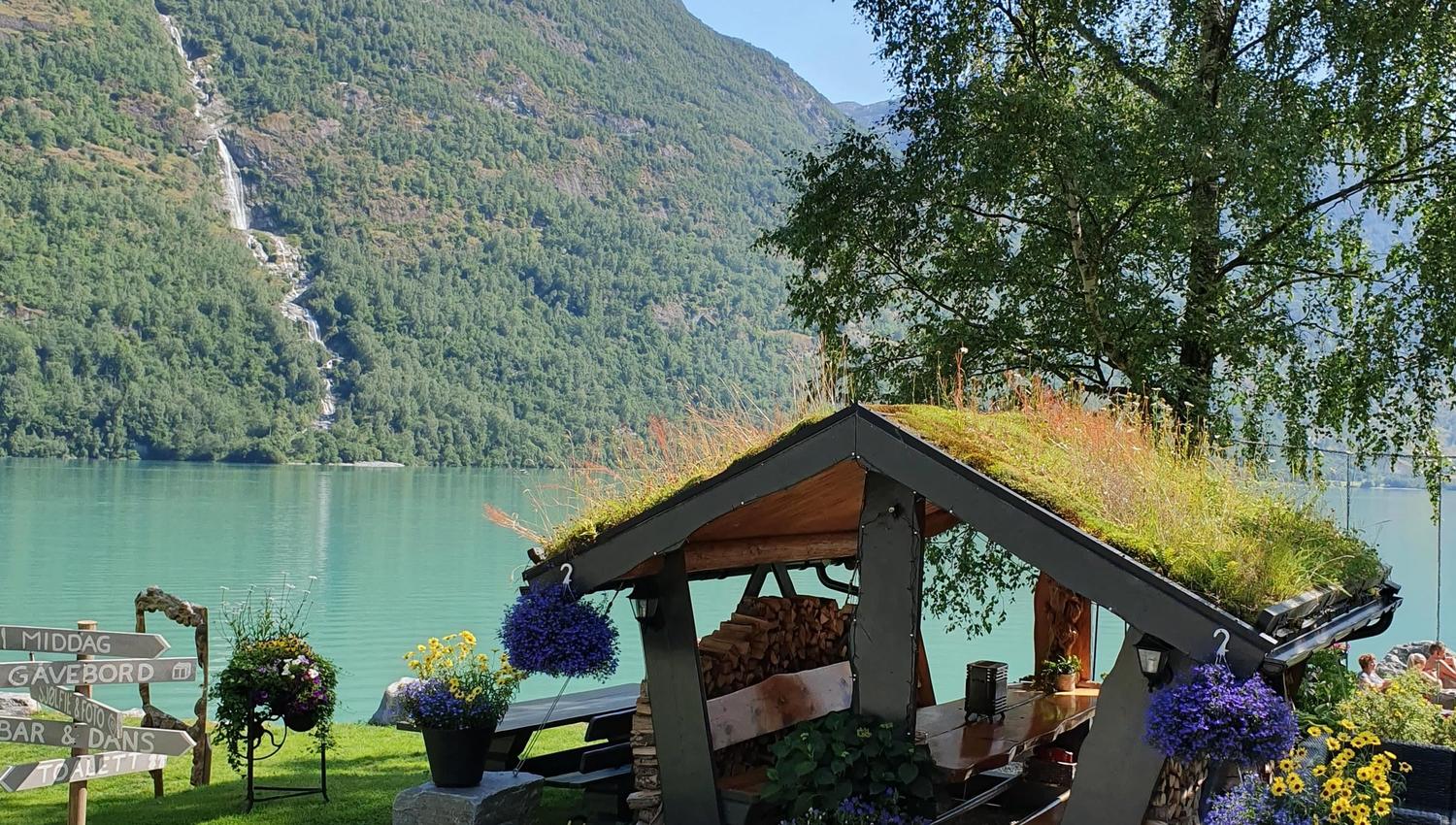 Small roofed shelter with potted flowers by a turquoise lake, with forested mountains and a waterfall in the distance..
