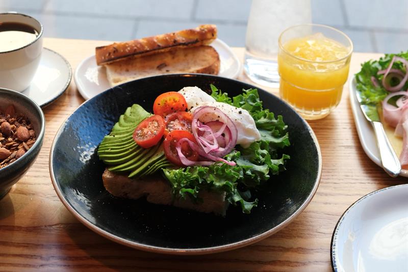 Detail of focaccia topped with avocado, tomato, red onion and lettuce, accompanied by a glass of orange juice, a cup of coffee, and a slice of bread o