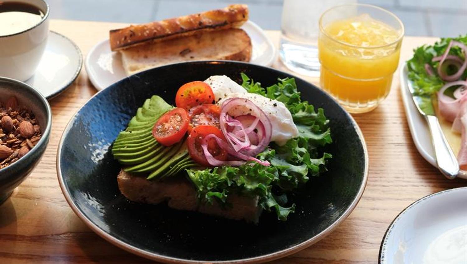 Detail of focaccia topped with avocado, tomato, red onion and lettuce, accompanied by a glass of orange juice, a cup of coffee, and a slice of bread o