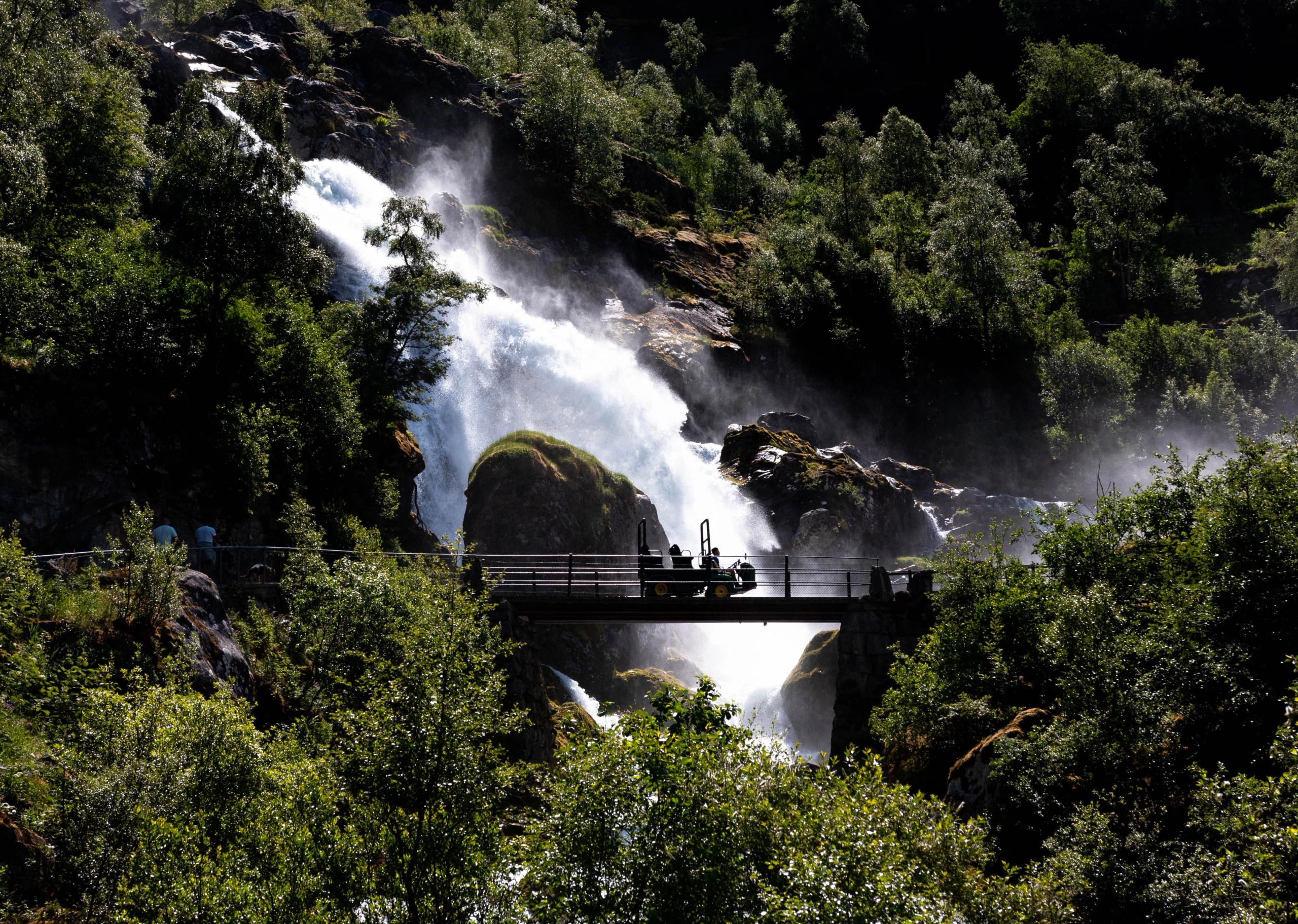 Sunlit waterfall cascades through rocky forest, with mist rising behind a small bridge and silhouettes of visitors..