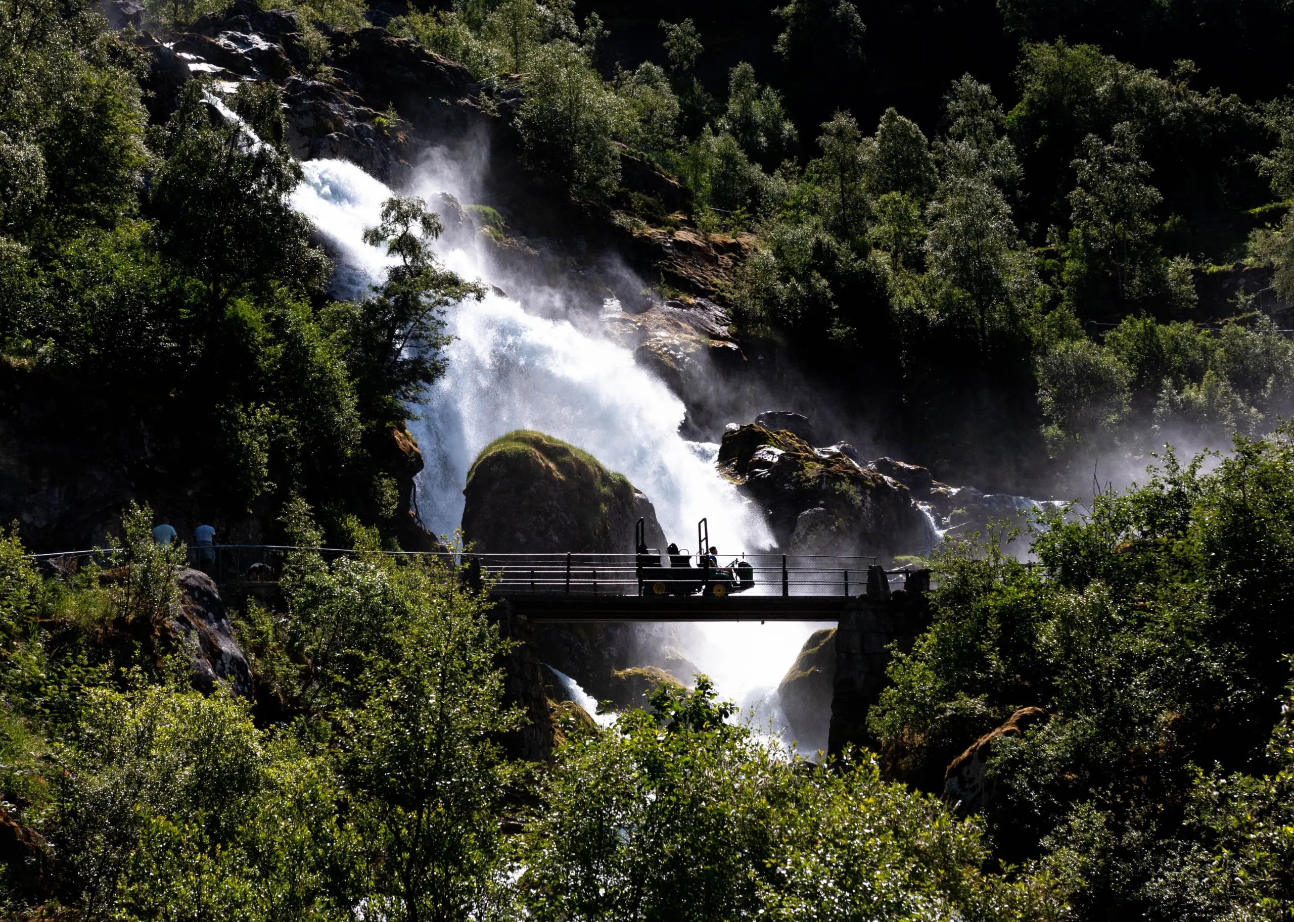 Sunlit waterfall cascades through rocky forest, with mist rising behind a small bridge and silhouettes of visitors..