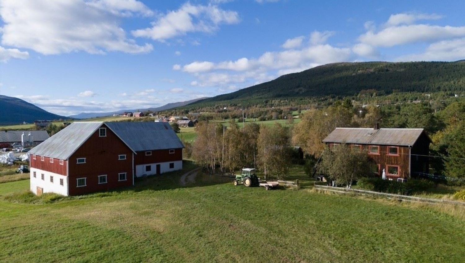 Aerial view of traditional farm buildings situated in an open valley with fields, trees, and mountains in the background under a clear sky.