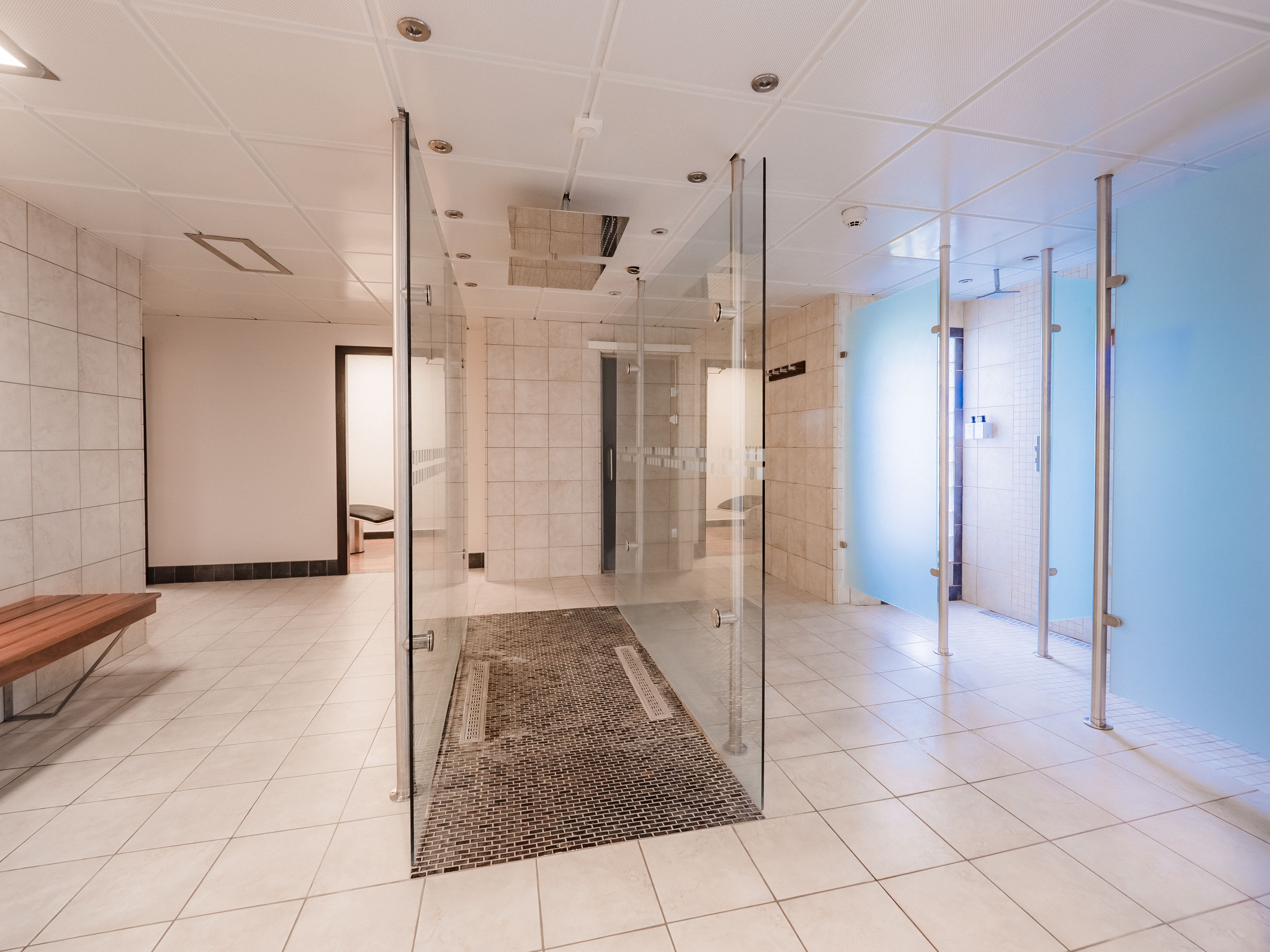 Empty tiled shower and locker room with glass partitions, ceiling fixtures, blue privacy panels, and a wooden bench.
