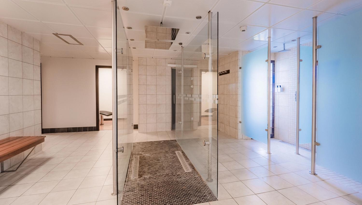 Empty tiled shower and locker room with glass partitions, ceiling fixtures, blue privacy panels, and a wooden bench.