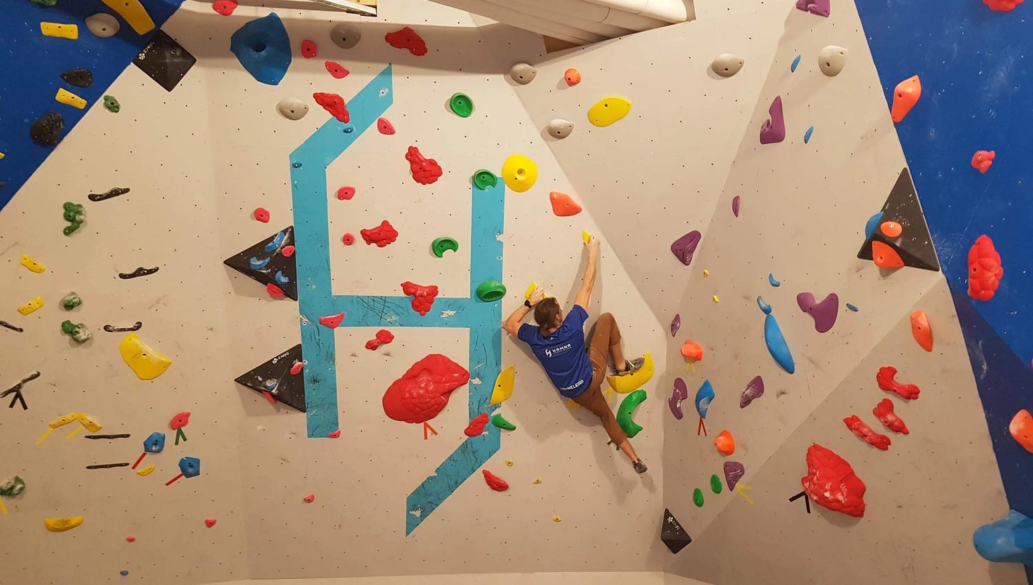 A climber in a blue shirt ascends an indoor bouldering wall with colorful holds on angled panels under ceiling lights..