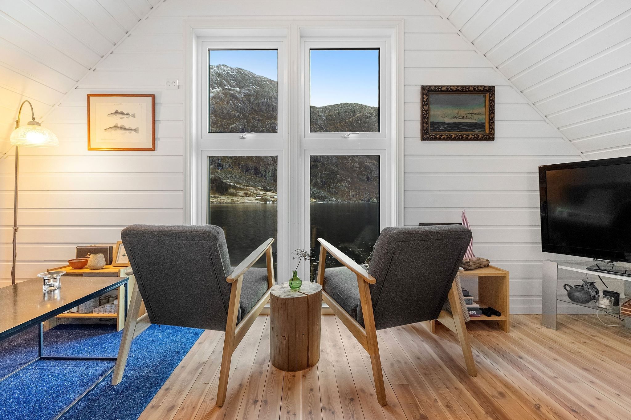 Two gray armchairs face tall windows overlooking a lake and rocky hills, in a white wood-paneled loft room at the Seahouse at Hornelen Panorama in Bremanger..