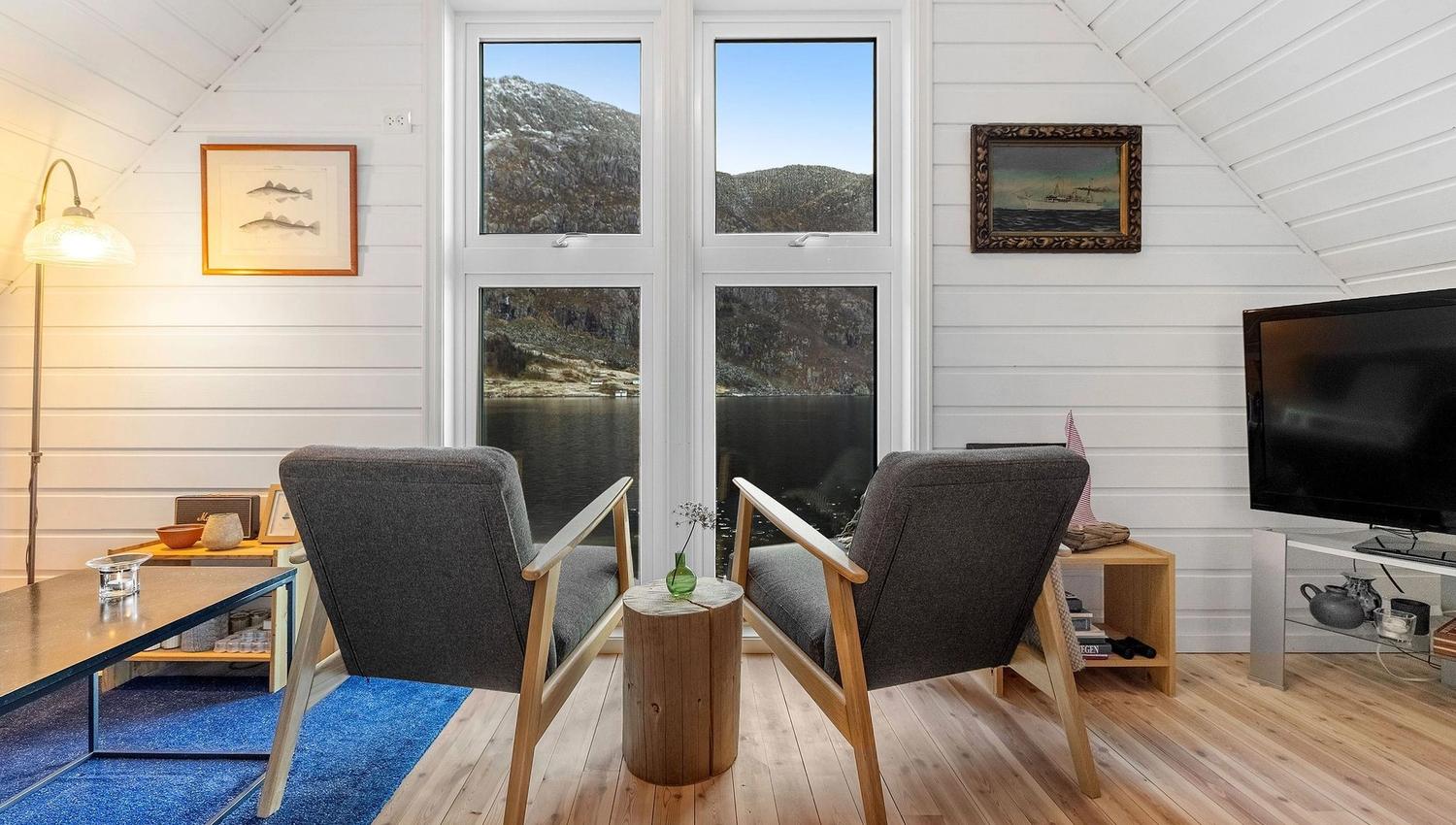 Two gray armchairs face tall windows overlooking a lake and rocky hills, in a white wood-paneled loft room at the Seahouse at Hornelen Panorama in Bremanger..
