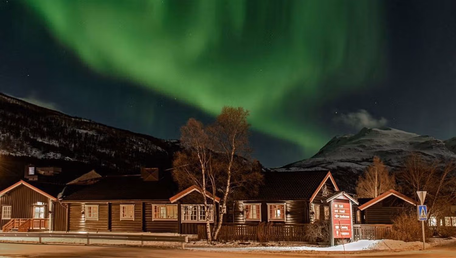 Northern lights over snow-covered guest lodge with mountains in the background at night