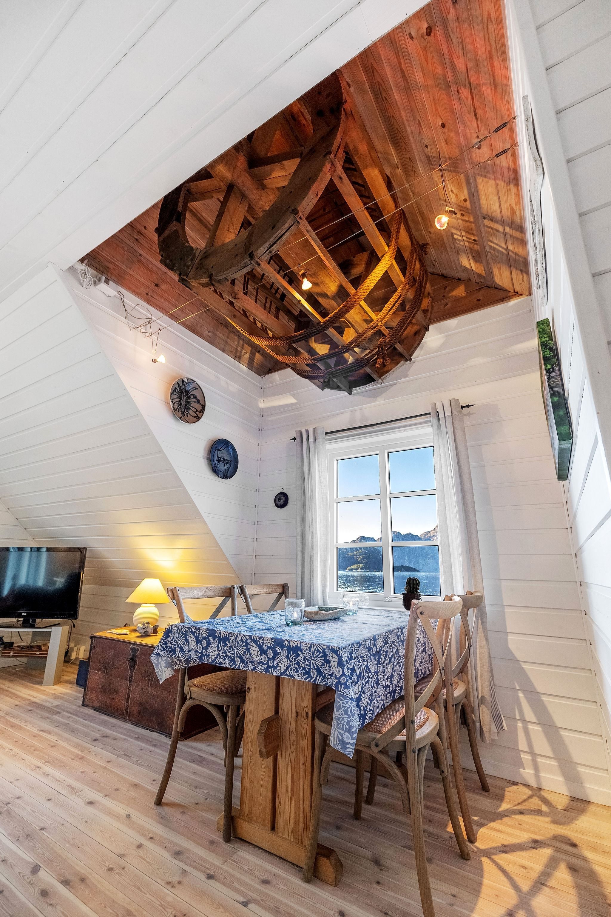 Bright white cabin dining area with wooden table and chairs, blue tablecloth, and rustic wooden ceiling feature above at the Seahouse at Hornelen Panorama in Bremanger..
