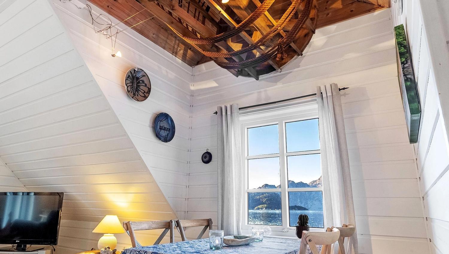 Bright white cabin dining area with wooden table and chairs, blue tablecloth, and rustic wooden ceiling feature above at the Seahouse at Hornelen Panorama in Bremanger..