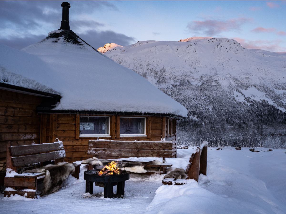 Snow-covered building and outdoor fire at Tromsø Lapland’s camp, surrounded by mountains and Arctic winter scenery.