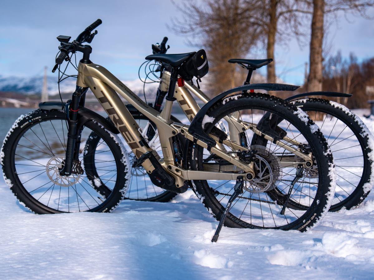 Two tan mountain bikes with thick tires stand in snow near a lake, with bare trees and distant hills behind.