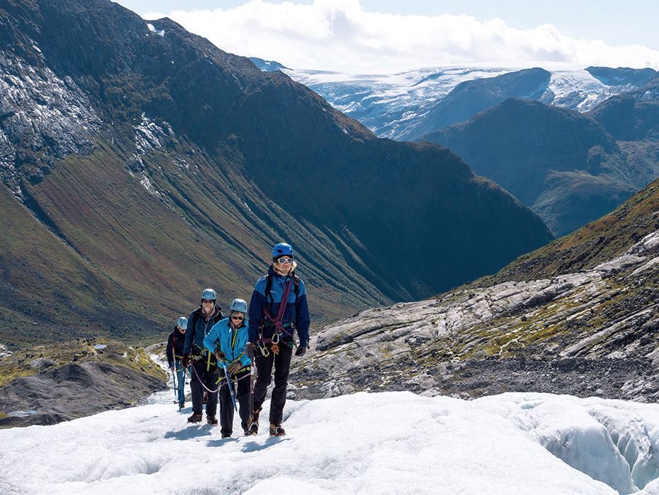 Four roped climbers in helmets and gear hike on a snowy glacier with steep mountains and a distant icefield behind..