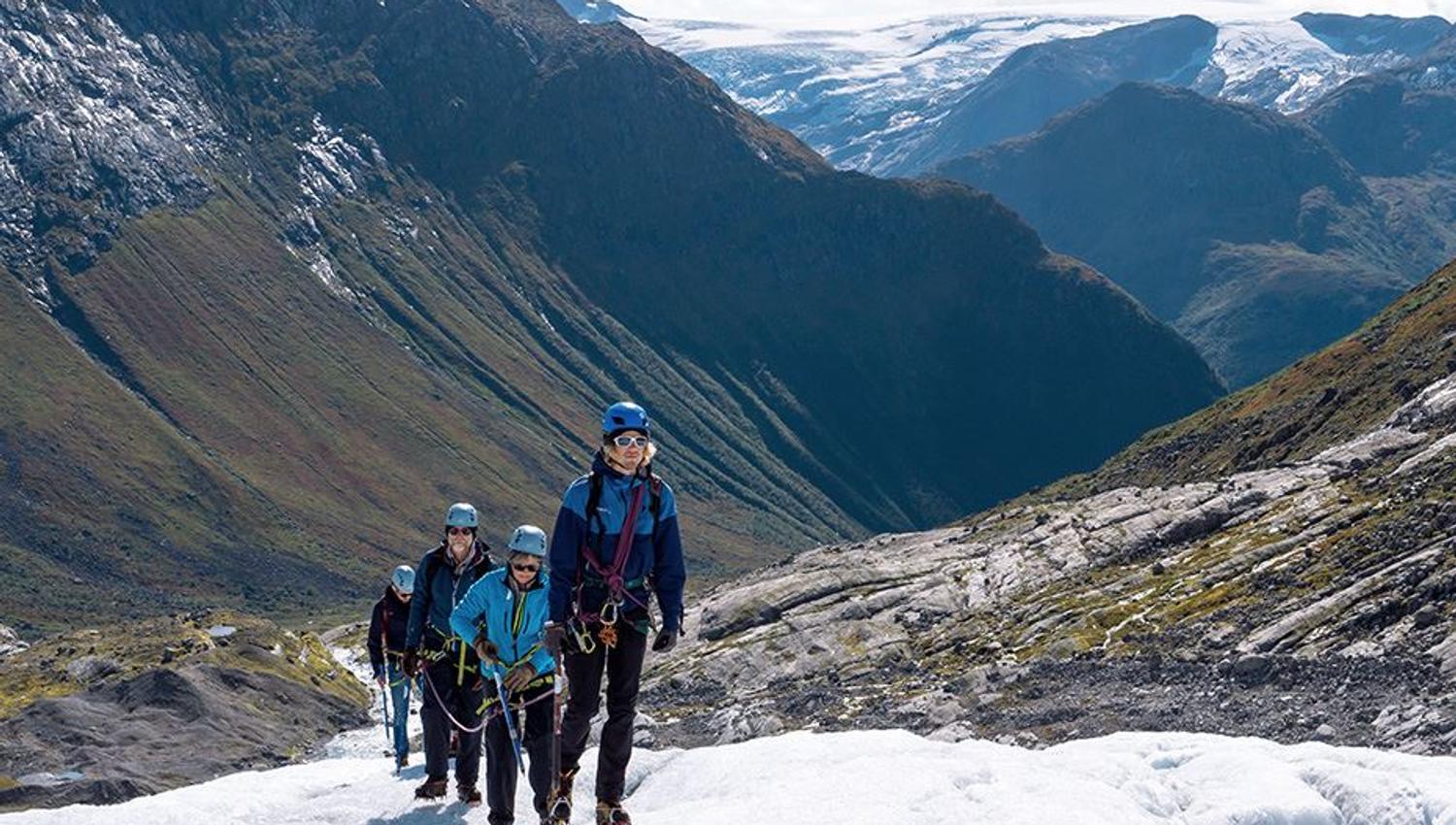 Four roped climbers in helmets and gear hike on a snowy glacier with steep mountains and a distant icefield behind..