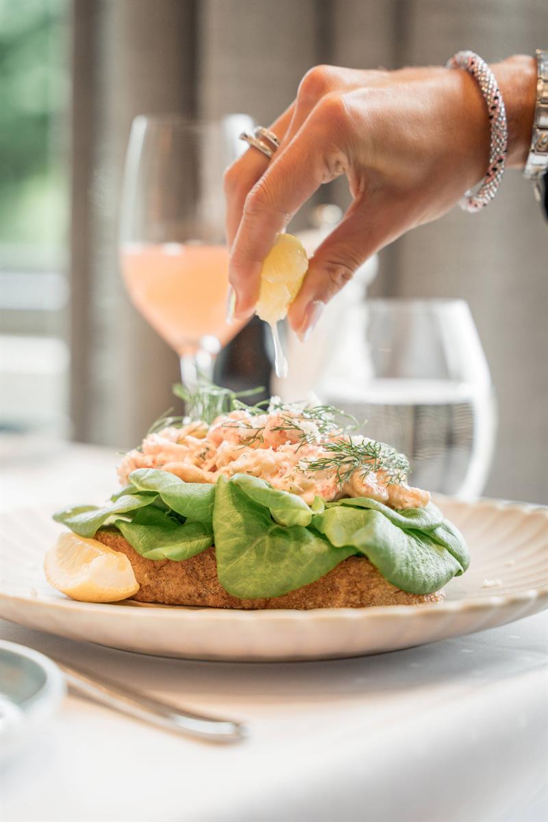 Hånd presser sitron over en rekesmørbrød toppet med friske urter og salatblader.