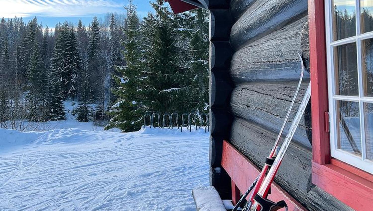 Svart og rød hytte i marka. Blå himmel og snø på bakken. Langrennsski står inntil hytteveggen.
