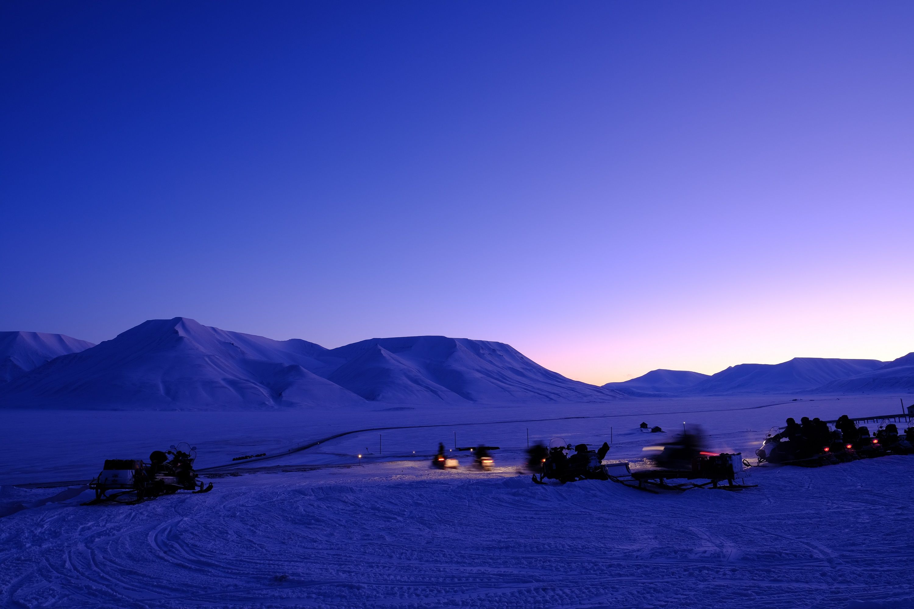 Persons on snowmobiles heading out on a trip with a mountainous landscape covered in blue light in the background