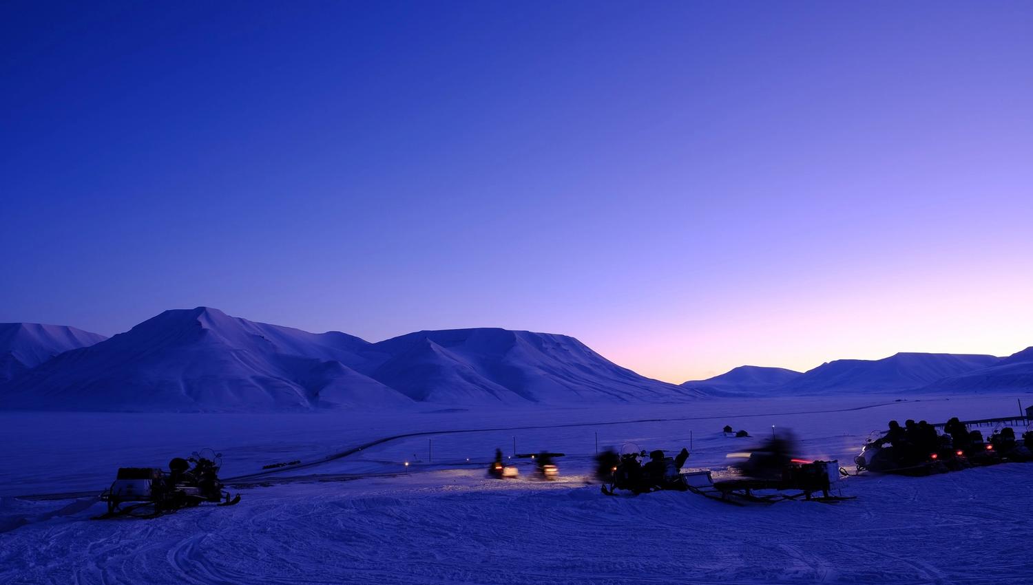 Persons on snowmobiles heading out on a trip with a mountainous landscape covered in blue light in the background