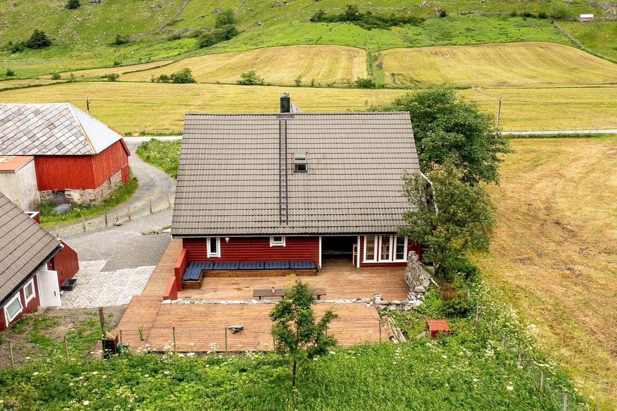 A red farmhouse with a gray roof and wooden deck sits beside a gravel drive, surrounded by green and golden fields  by Hoddevik Surf House at Stadlandet..