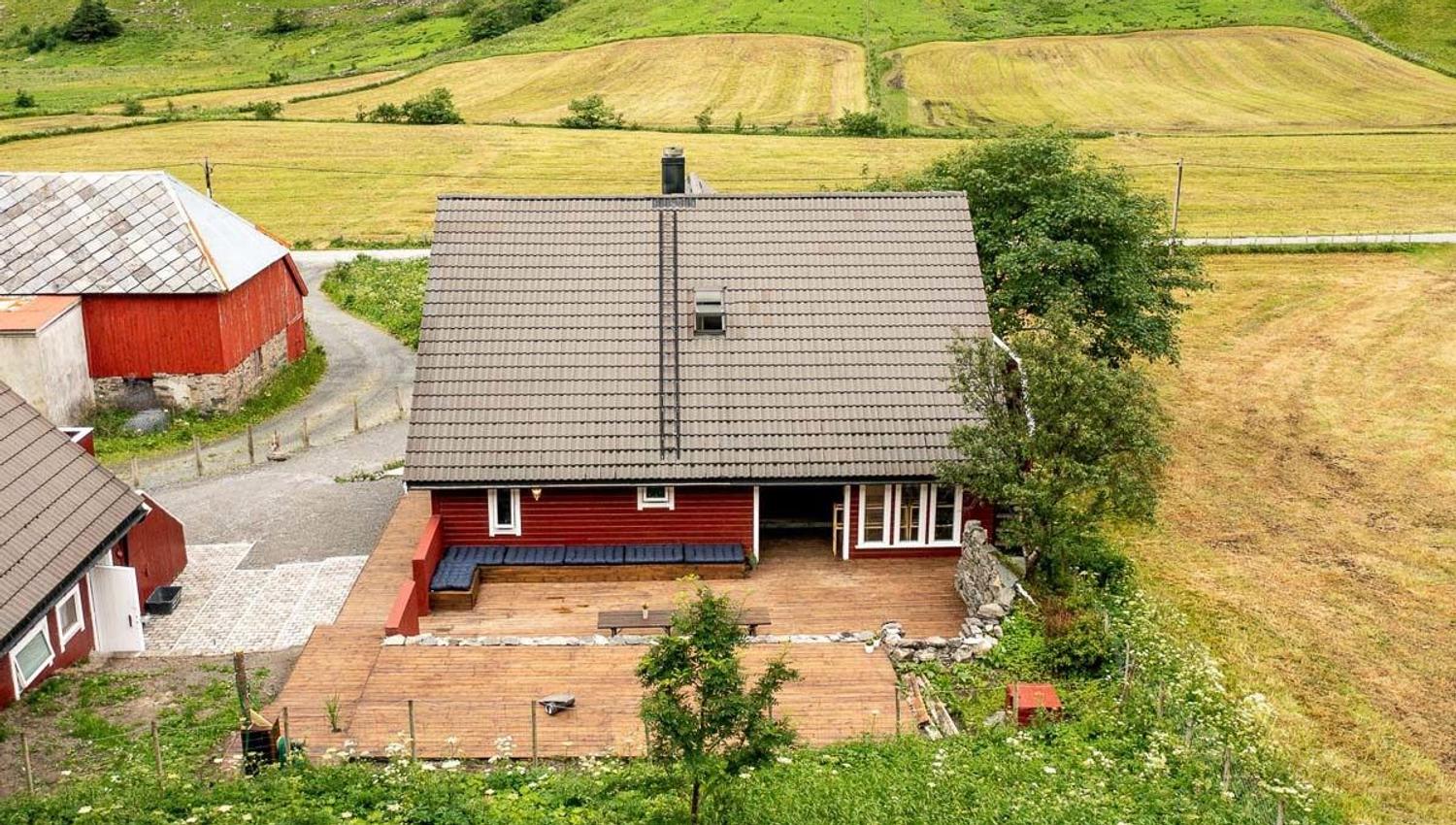 A red farmhouse with a gray roof and wooden deck sits beside a gravel drive, surrounded by green and golden fields by Hoddevik Surf House at Stadlandet..