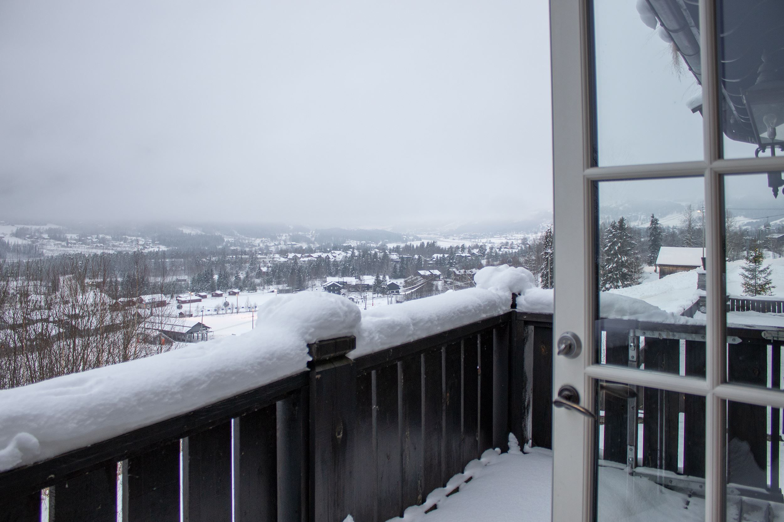 Utsikt från en balkong med svart räcke täckt av snö, mot ett vinterlandskap med hus, träd och dimmiga berg.