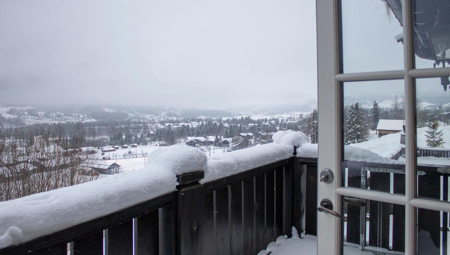 Utsikt från en balkong med svart räcke täckt av snö, mot ett vinterlandskap med hus, träd och dimmiga berg.
