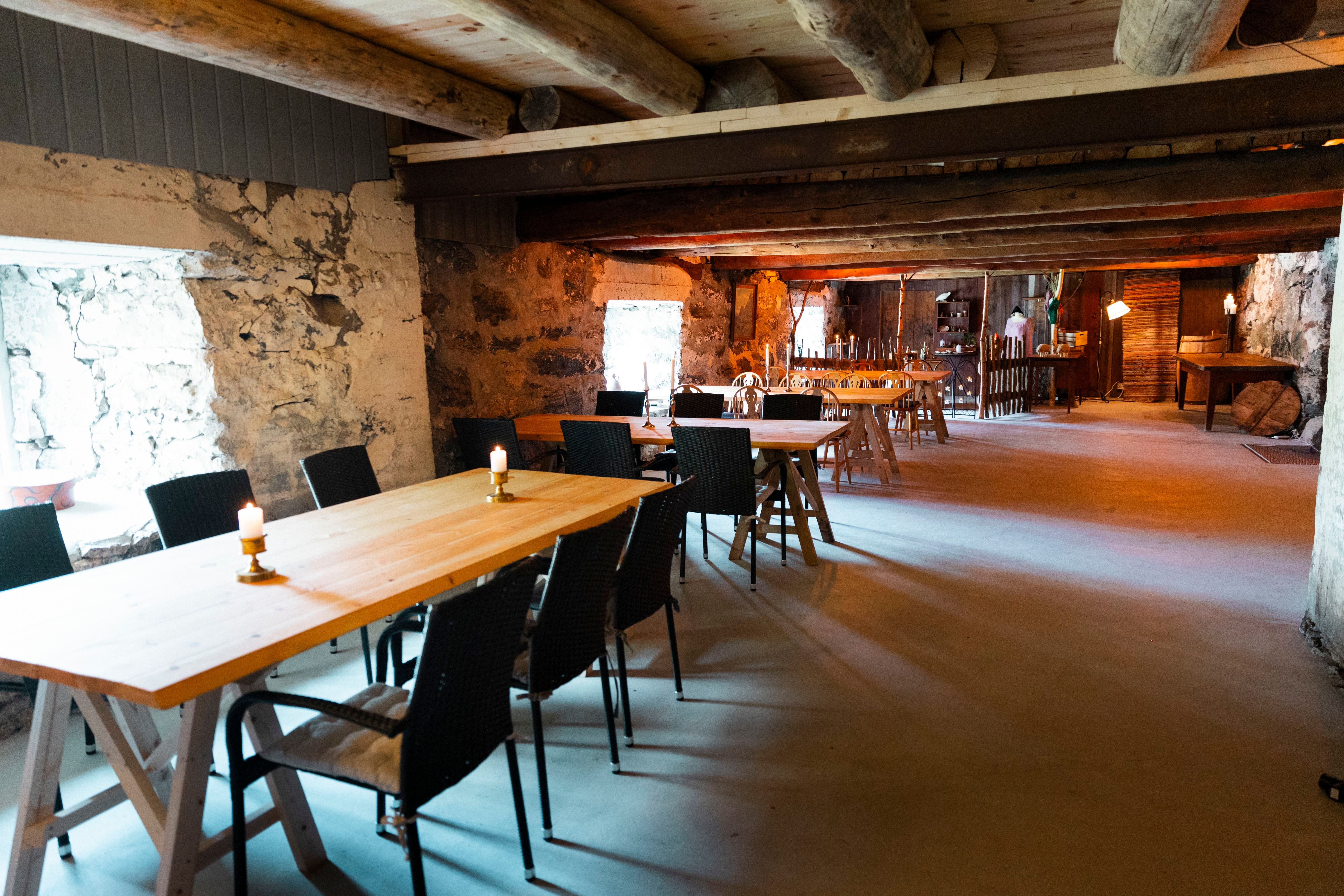 Long rustic room with wooden tables and black chairs, stone walls, exposed beams, candles, and warm lights.