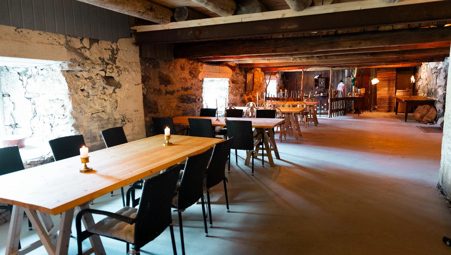 Long rustic room with wooden tables and black chairs, stone walls, exposed beams, candles, and warm lights.