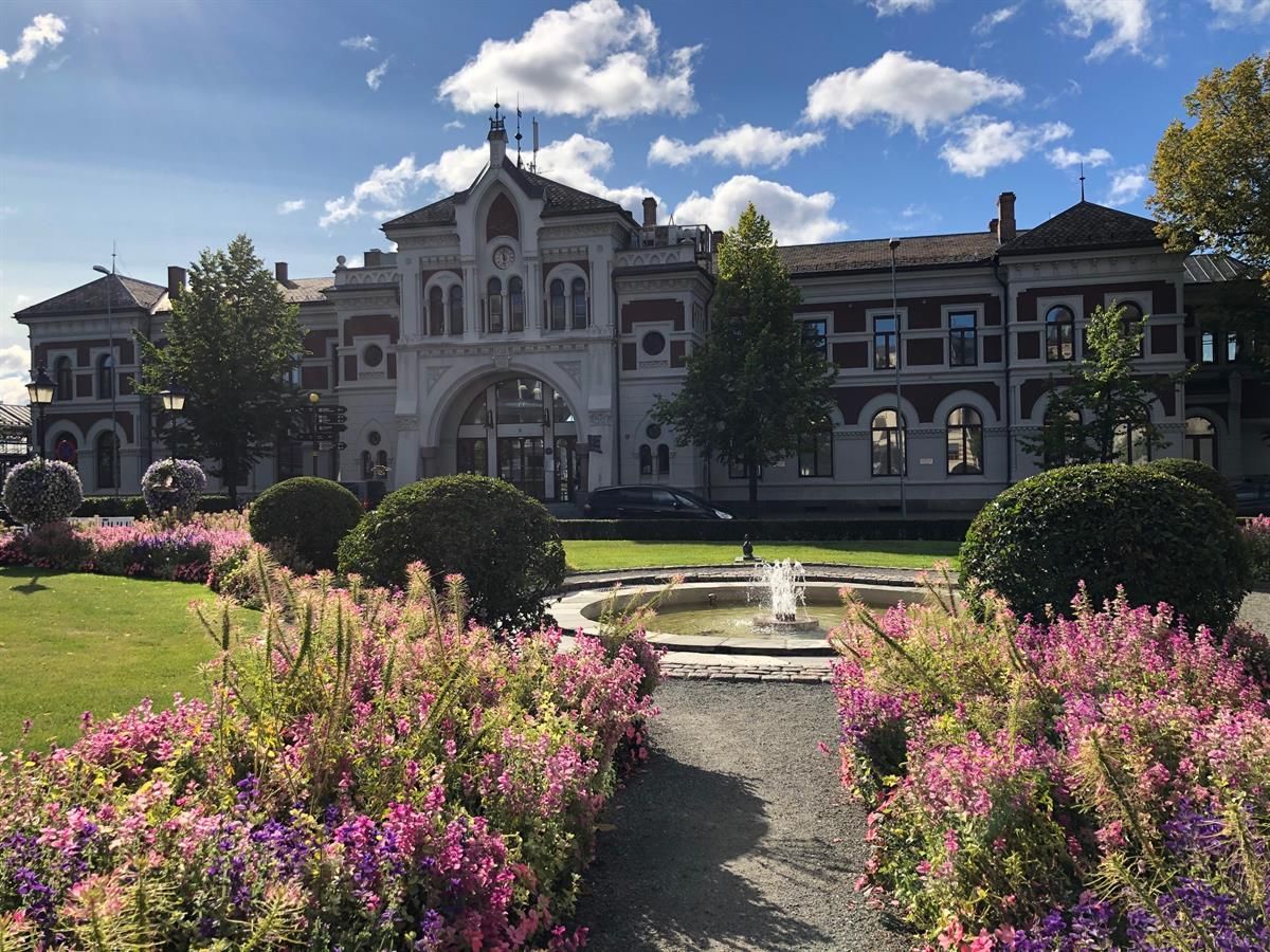 A sunny view of Jernbaneparken in Hamar, featuring colorful flowerbeds and a fountain in the foreground. In the background stands the historic Hamar S