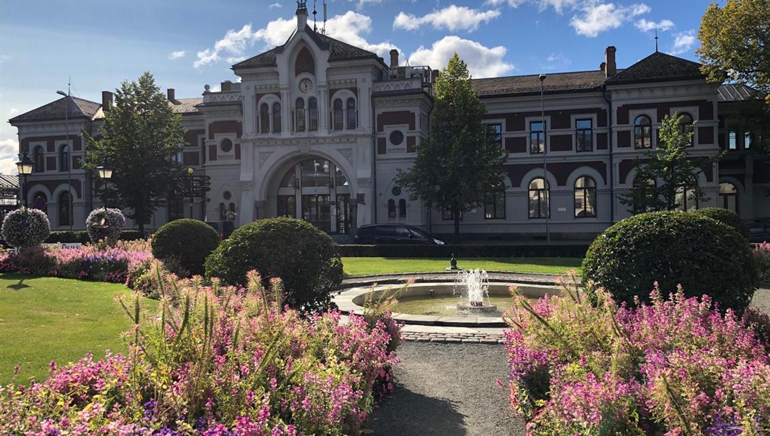 A sunny view of Jernbaneparken in Hamar, featuring colorful flowerbeds and a fountain in the foreground. In the background stands the historic Hamar S