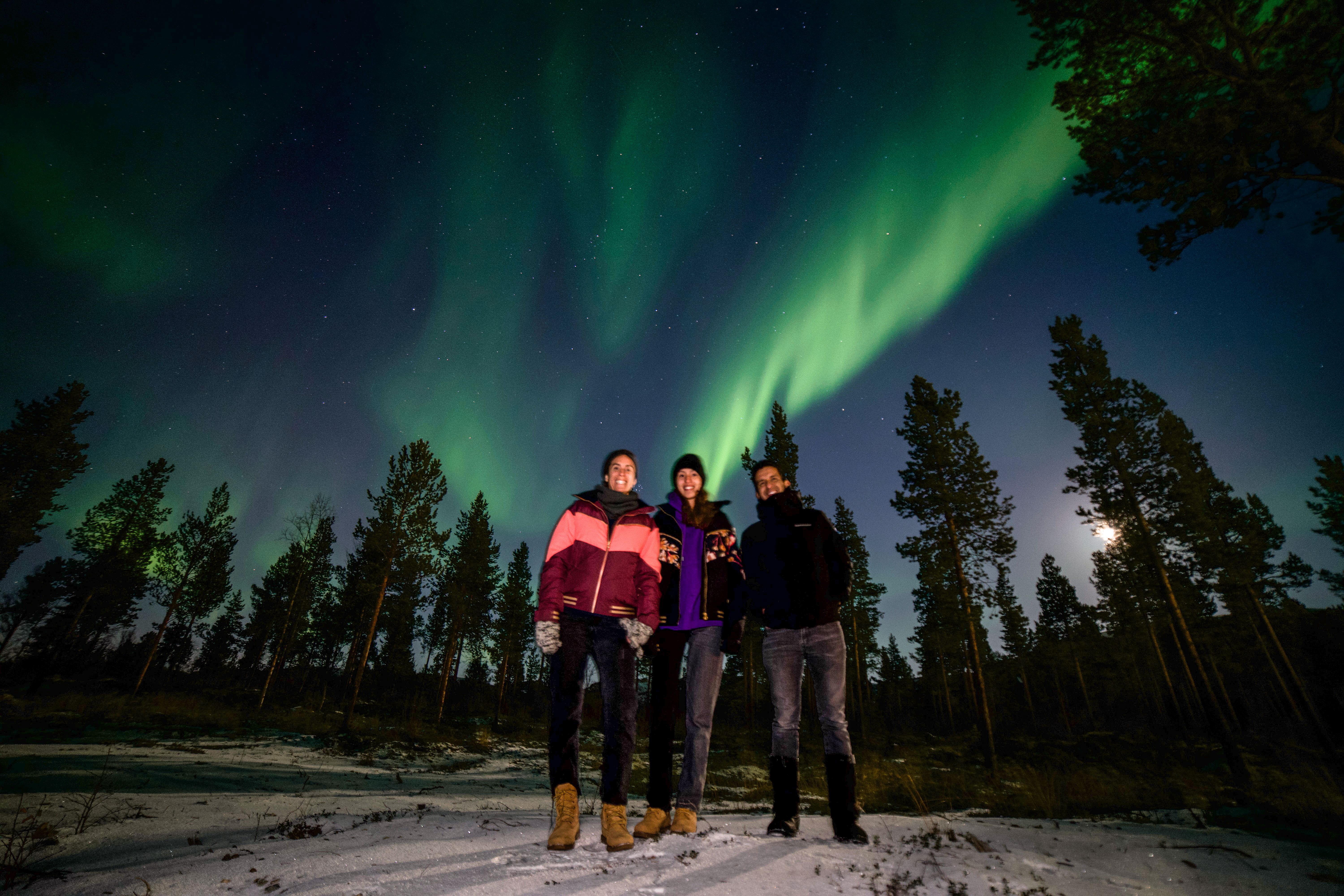 Witness the magic of the aurora borealis! Three friends capture a moment against the mesmerizing display of green lights dancing in the night sky, surrounded by a serene winter landscape. An unforgettable experience in nature!