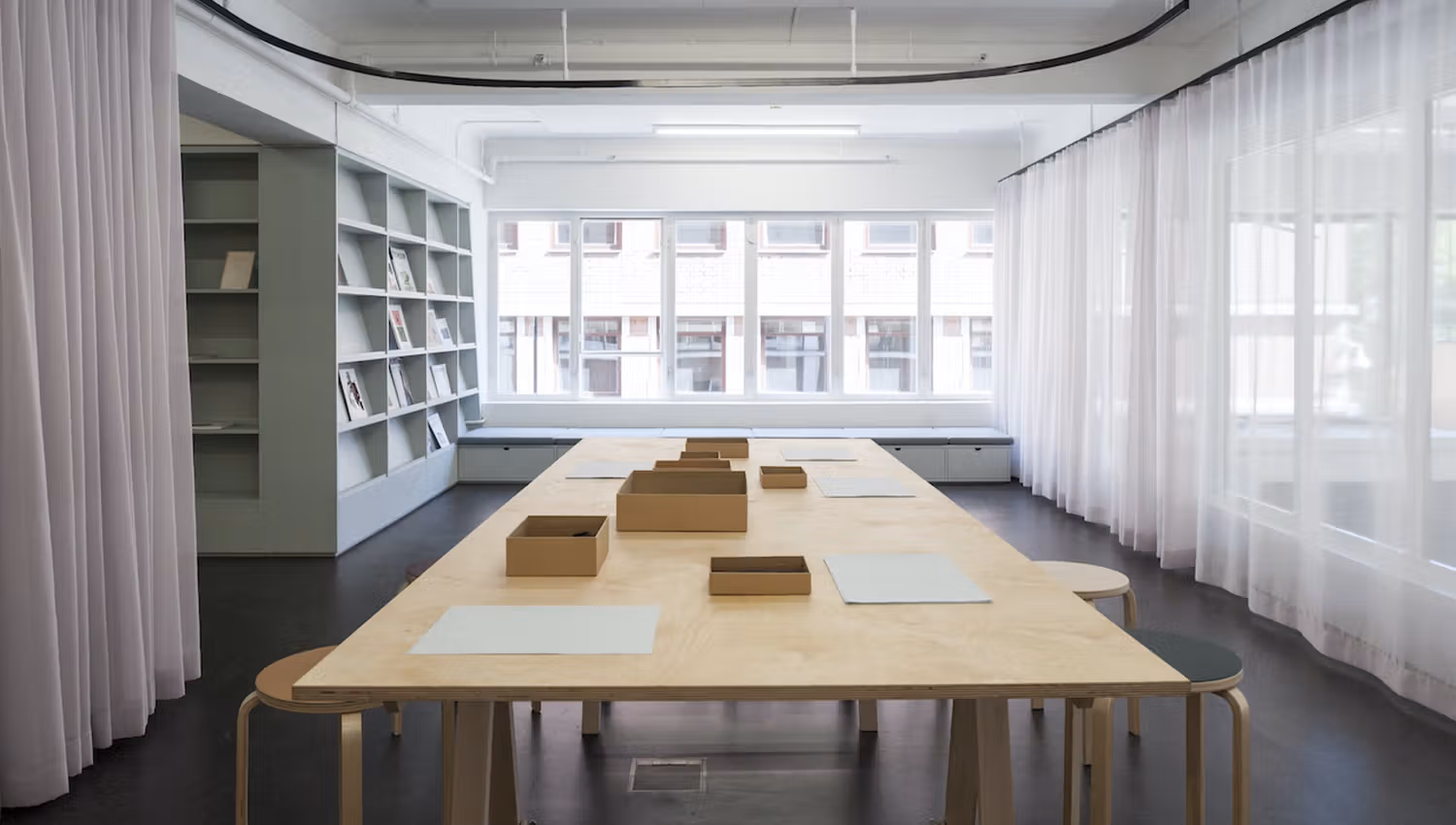 Conference room with a large light wooden table, stools, white curtains, bookshelves and a window.