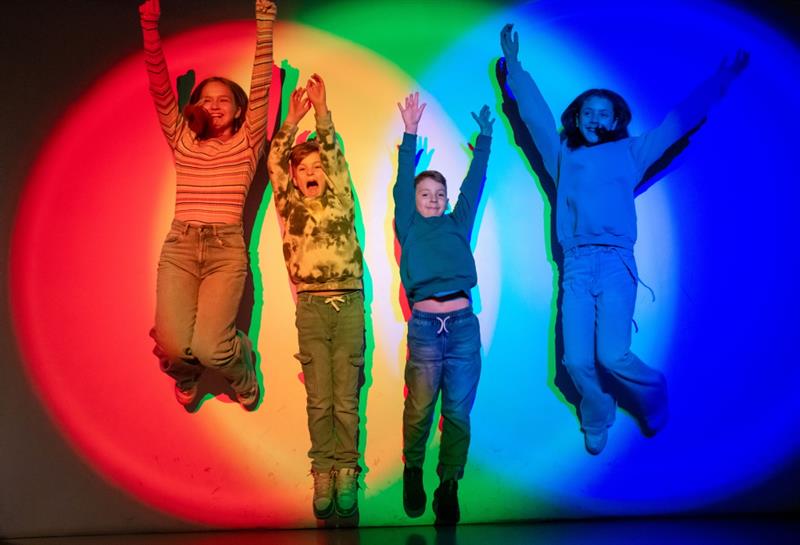 Four children jump in front of a brightly coloured wall, casting vibrant multi-coloured shadows behind them