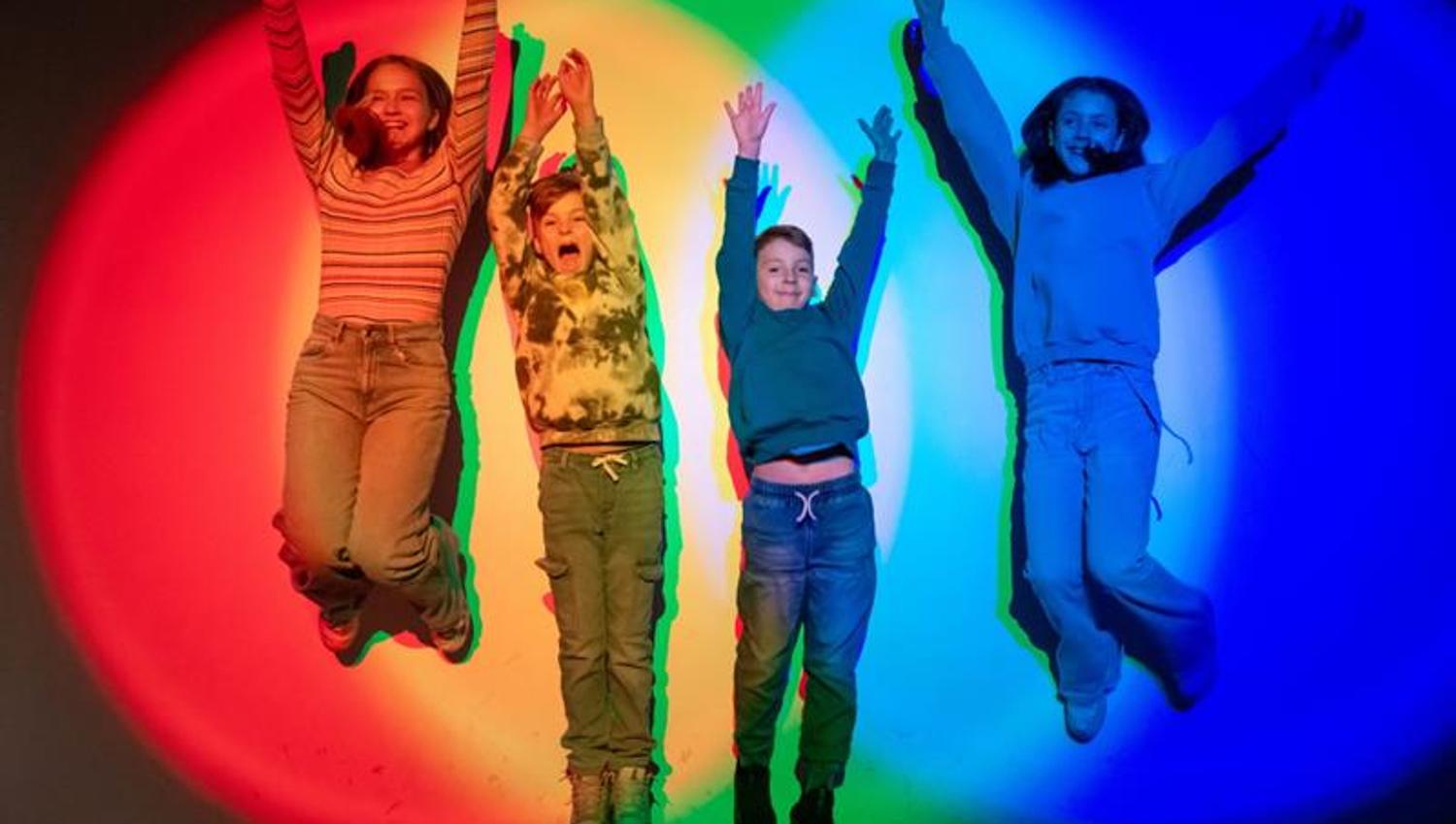 Four children jump in front of a brightly coloured wall, casting vibrant multi-coloured shadows behind them