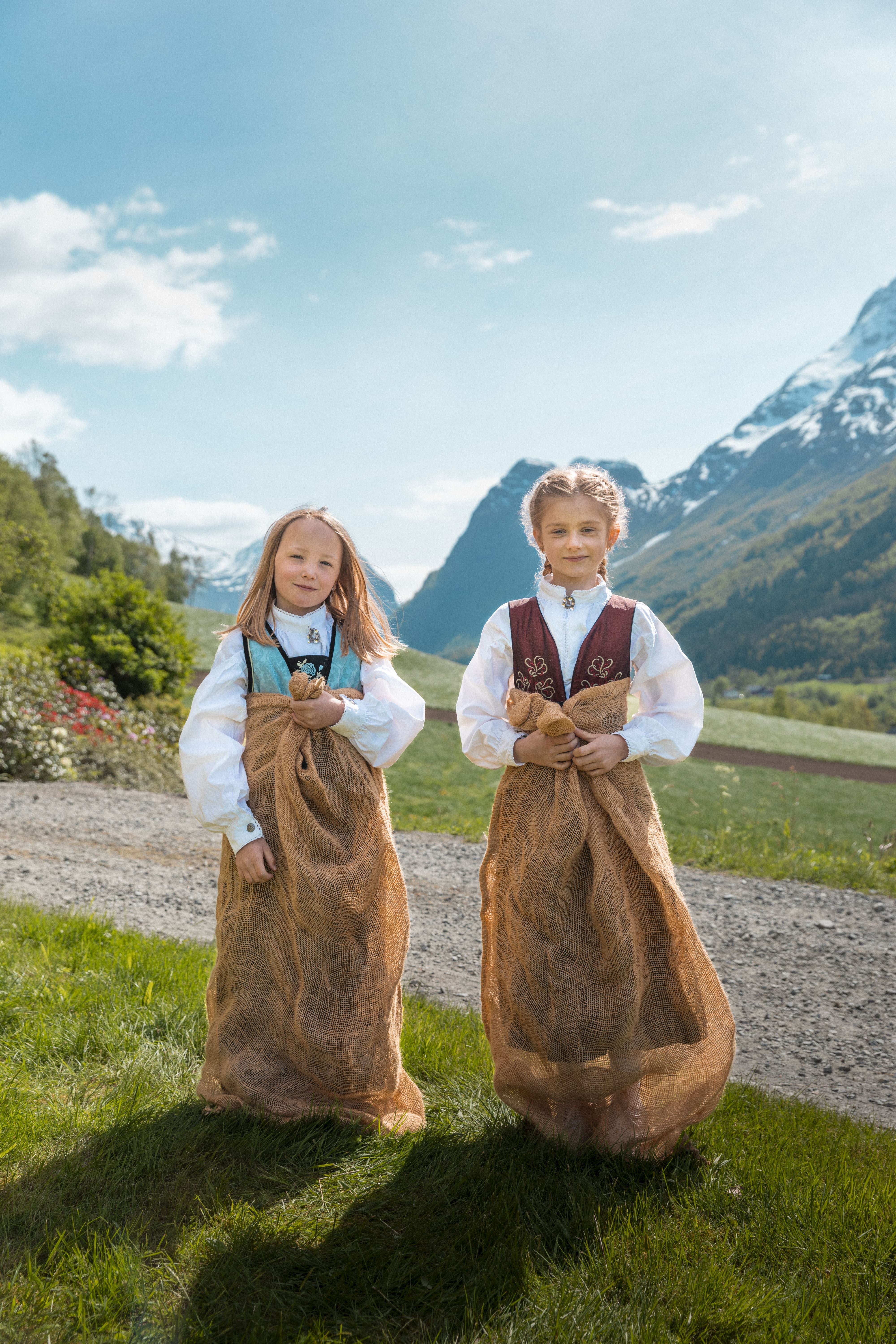 Two children in traditional outfits stand in burlap sacks on grass, with mountains, a path, and blue sky behind.