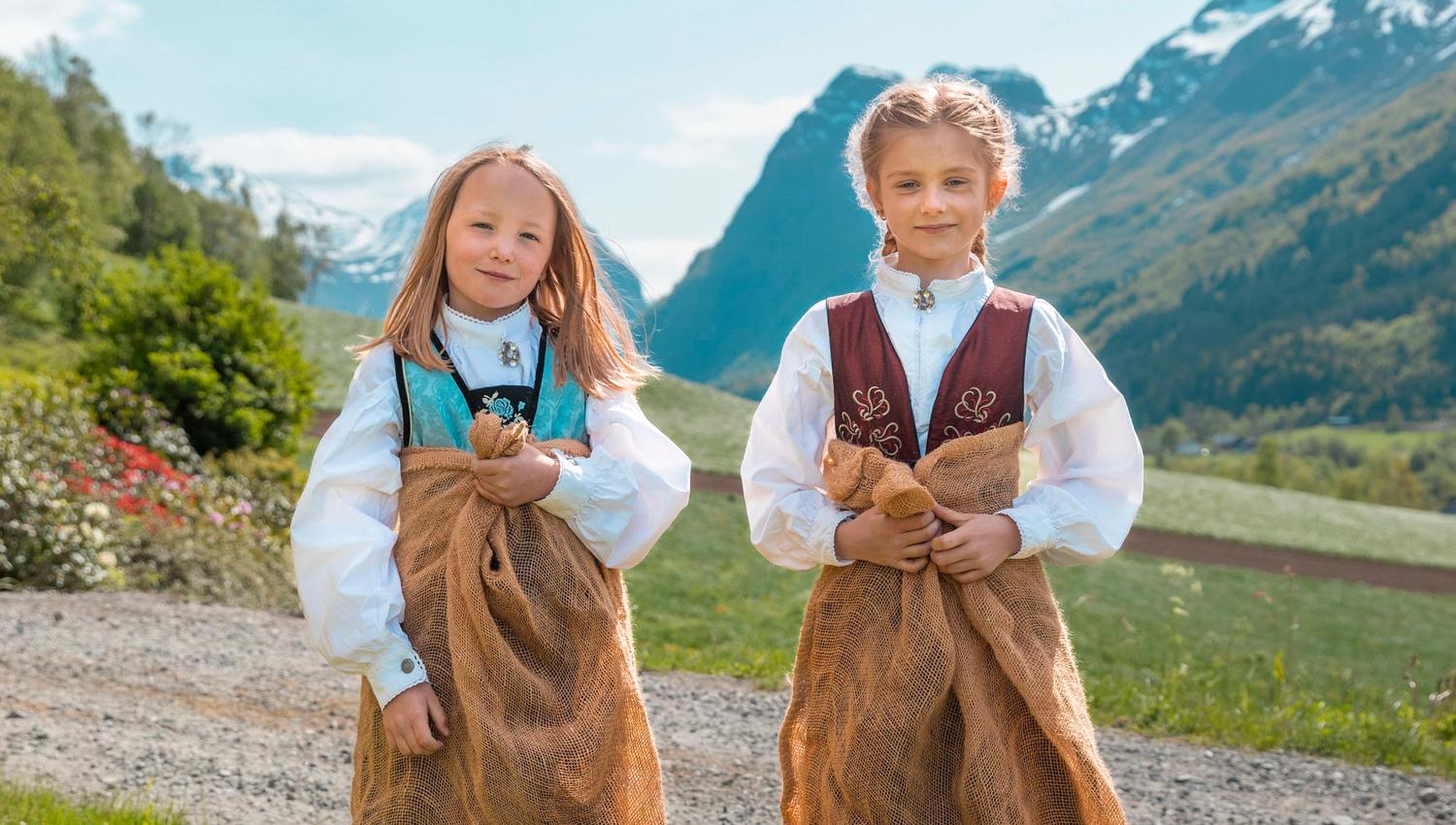 Two children in traditional outfits stand in burlap sacks on grass, with mountains, a path, and blue sky behind.