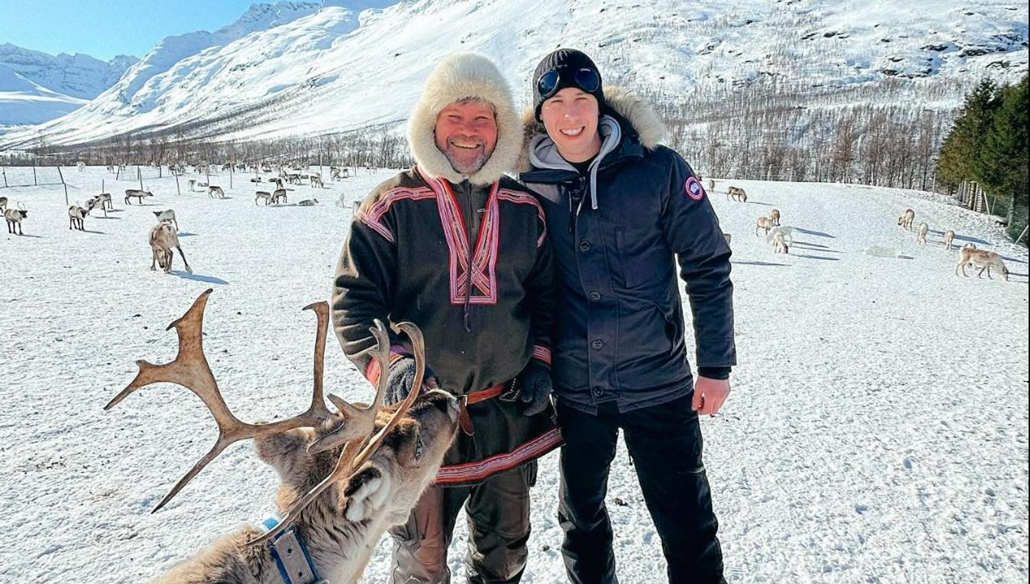 A Sami guide standing with a guest while a reindeer is looking at them