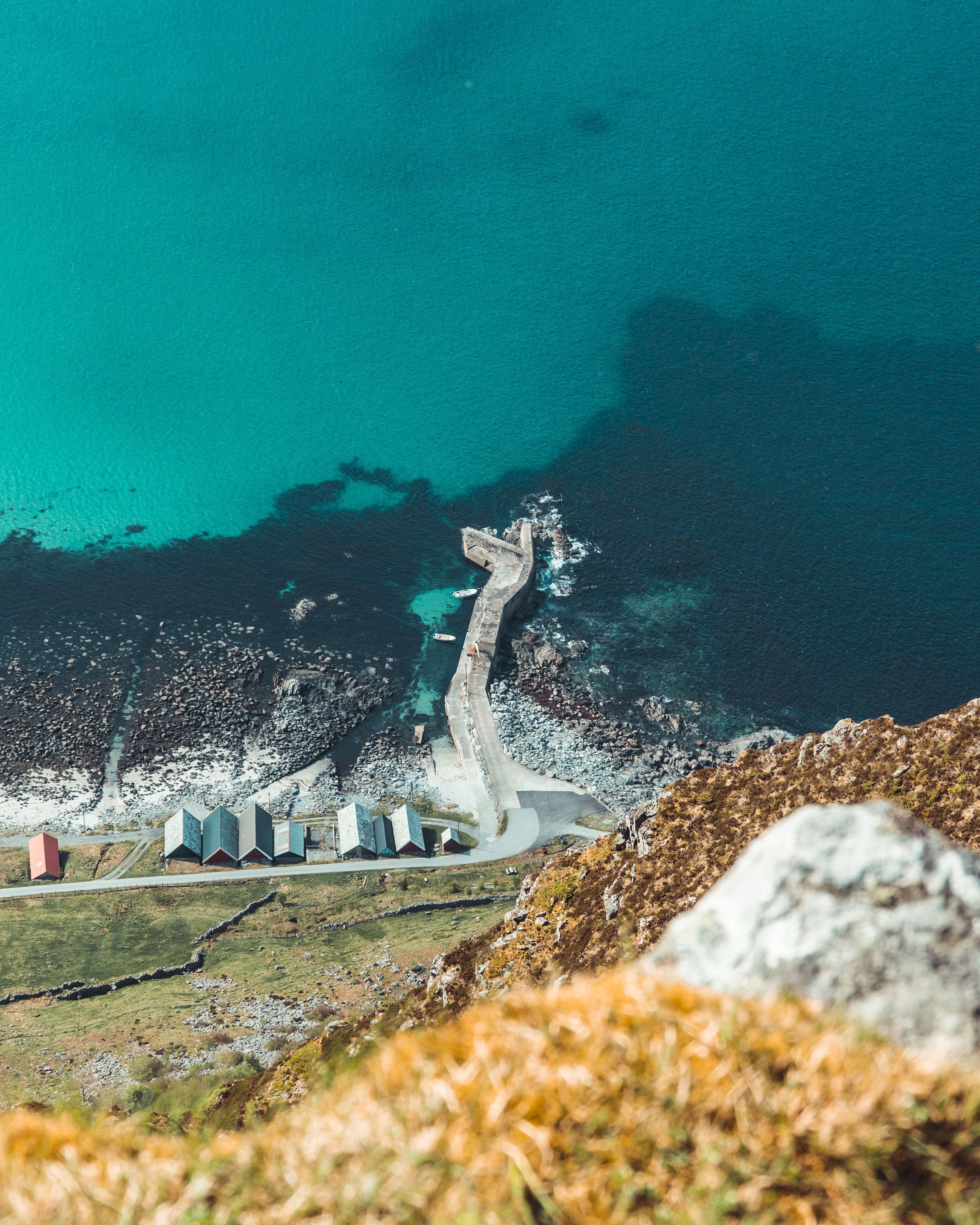 Utsyn frå ein høgde ned mot ein steinmolo, sjøbuer og steinstrand ved turkis hav og klippekyst..