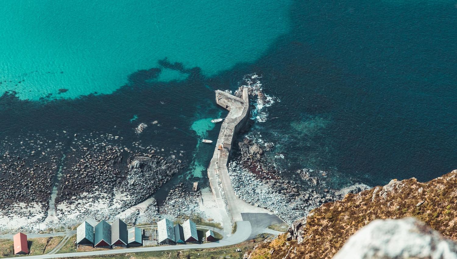 Utsyn frå ein høgde ned mot ein steinmolo, sjøbuer og steinstrand ved turkis hav og klippekyst..