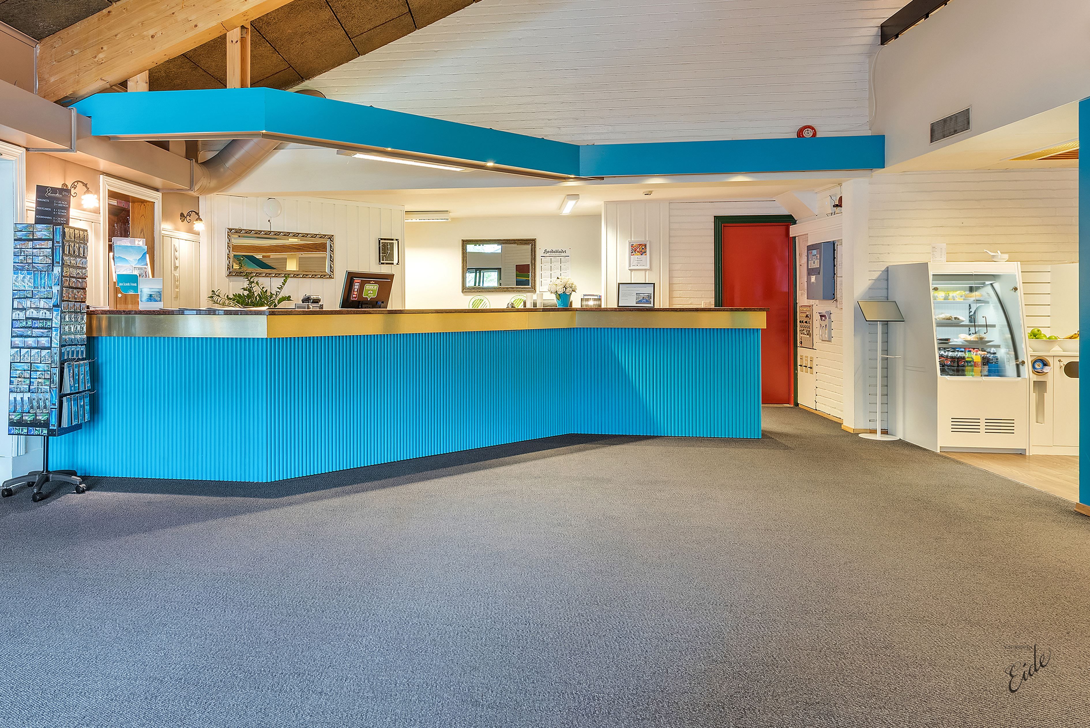 Empty hotel reception area with a long blue front desk, white walls, a red door, and a snack cooler on the right.