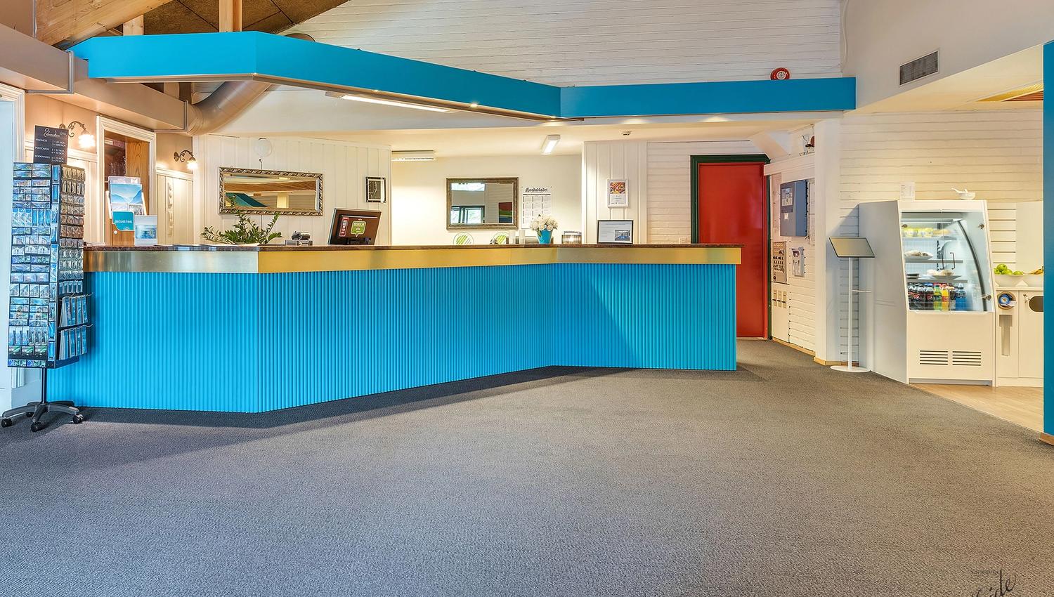 Empty hotel reception area with a long blue front desk, white walls, a red door, and a snack cooler on the right.