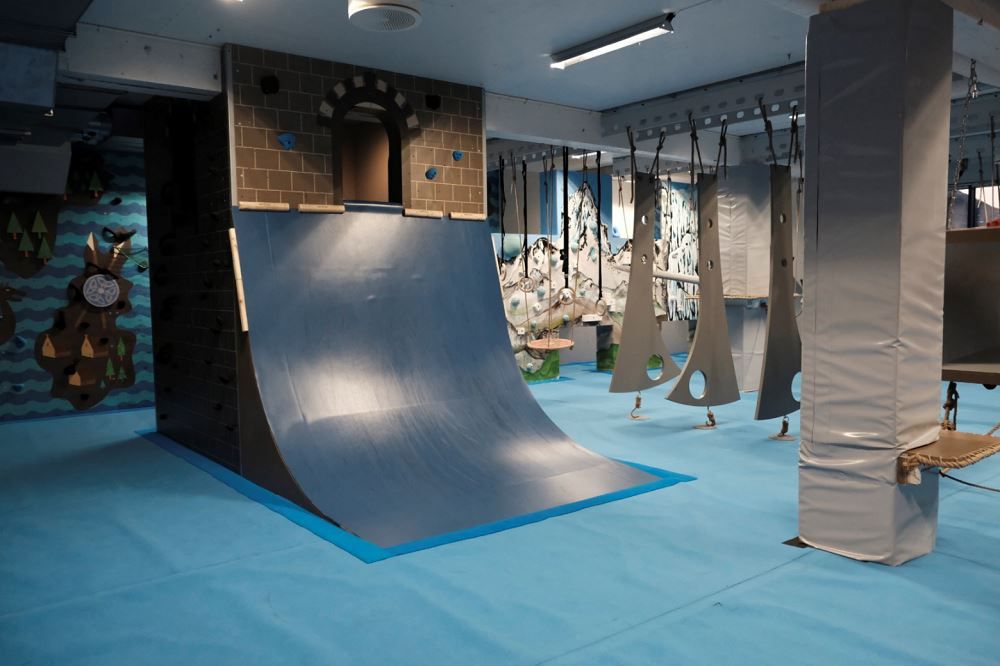 Indoor adventure gym with a curved ramp wall, hanging obstacles, padded columns, and blue safety flooring..