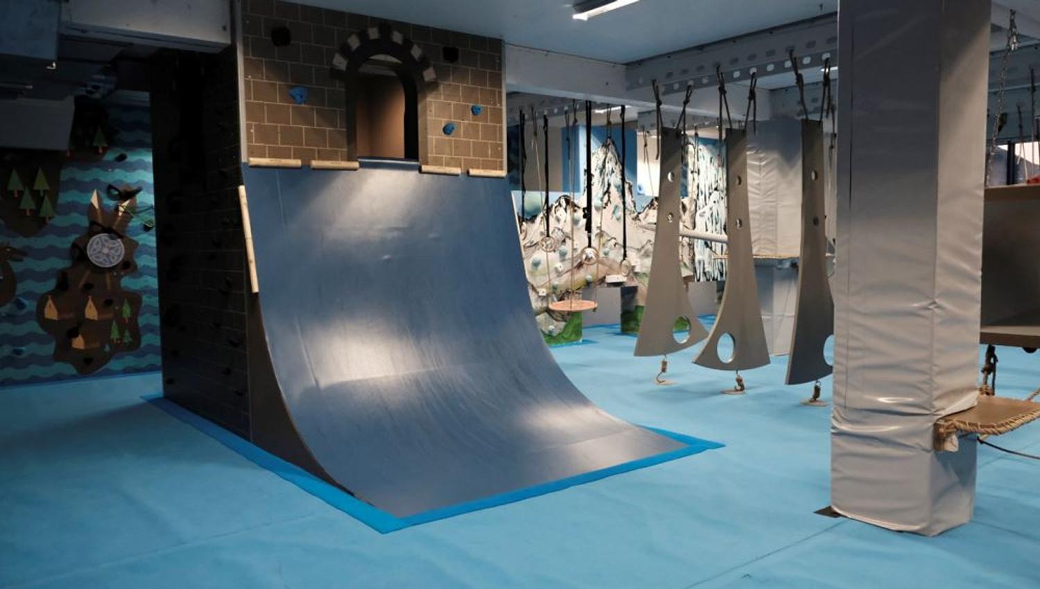 Indoor adventure gym with a curved ramp wall, hanging obstacles, padded columns, and blue safety flooring..