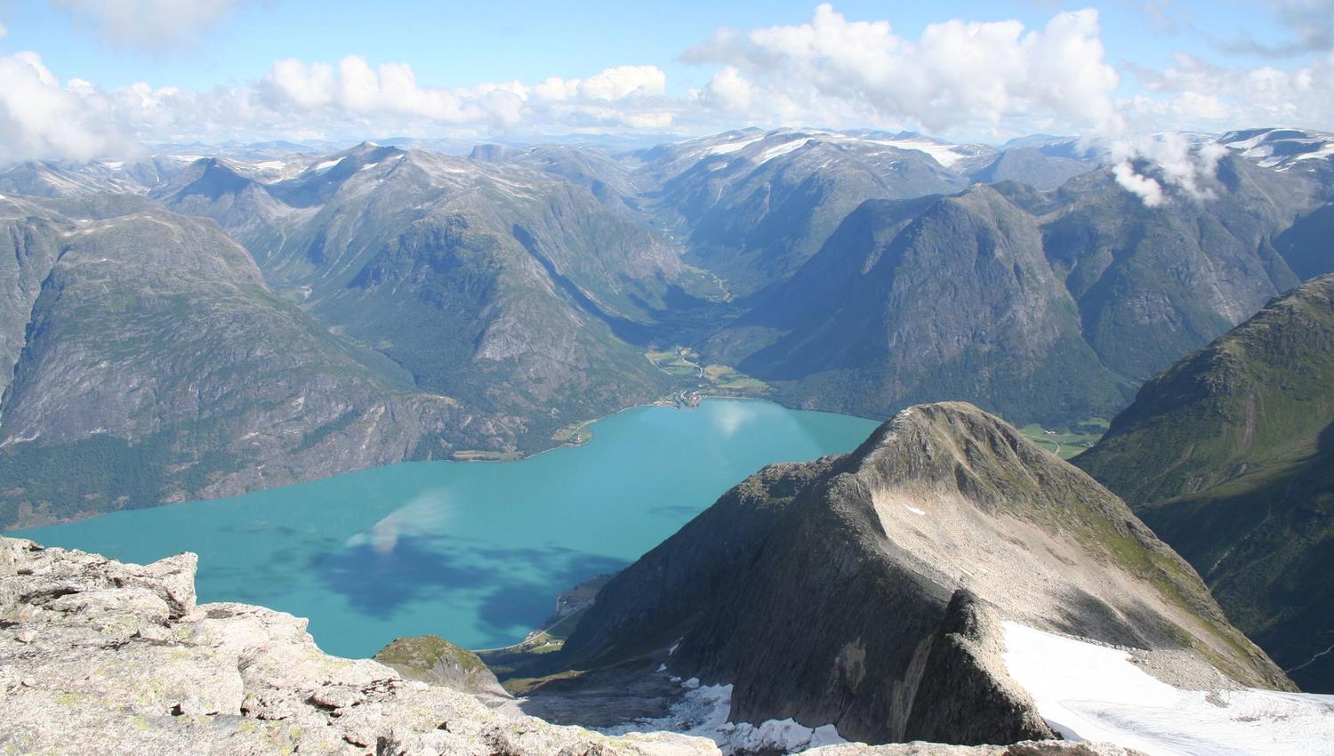 High-altitude view of a turquoise lake between steep mountain peaks, with rocky foreground and scattered clouds.