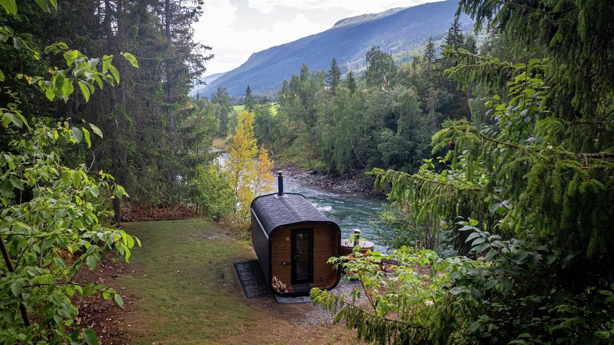 Wood-fired sauna located at the edge of a forest beside a river, with valley and mountains in the background.
